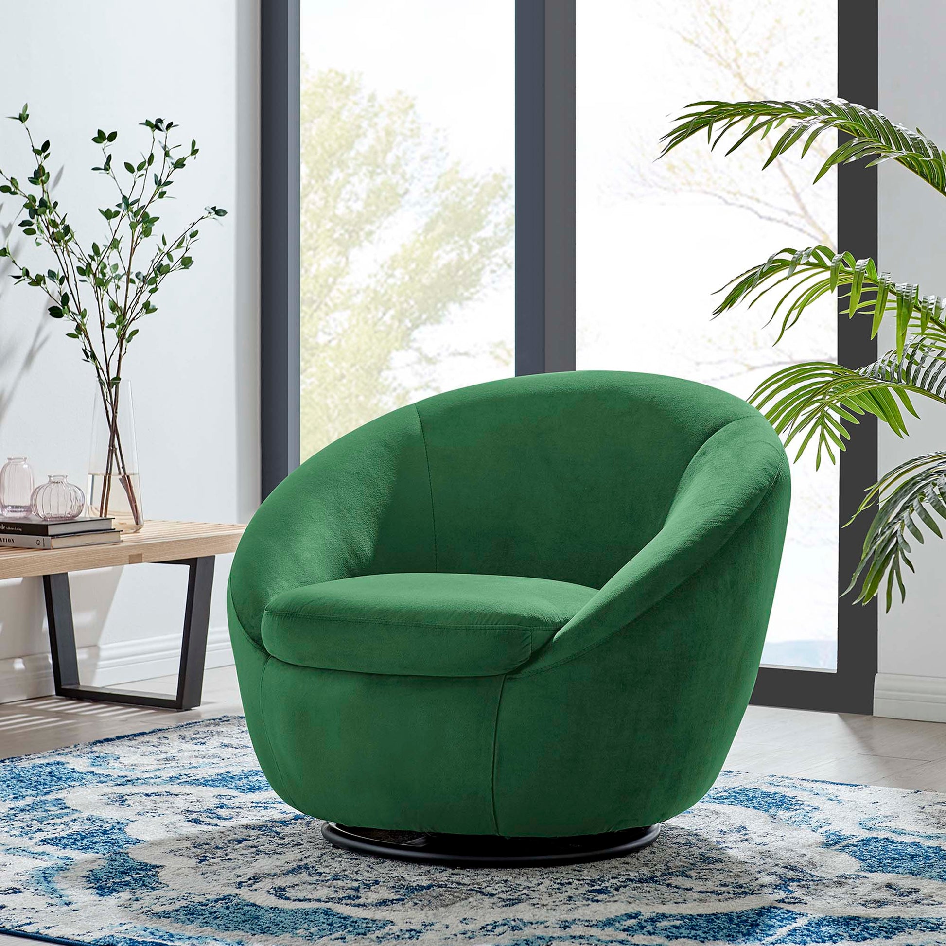 Emerald green velvet round swivel chair on a black pedestal base in a modern living room with a blue rug and large windows.