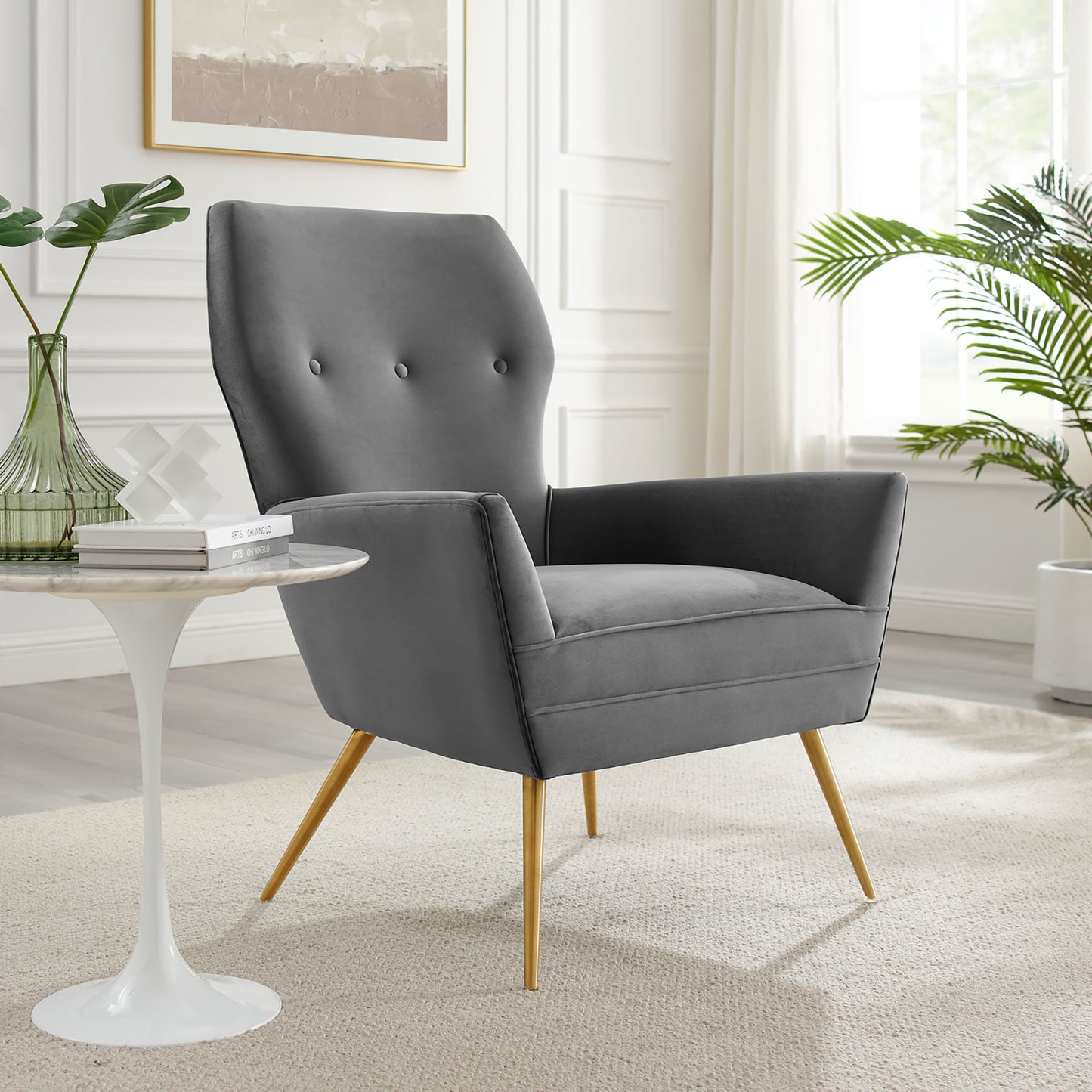 Gray velvet mid-century armchair with button-tufted back and gold tapered legs in a bright living room.