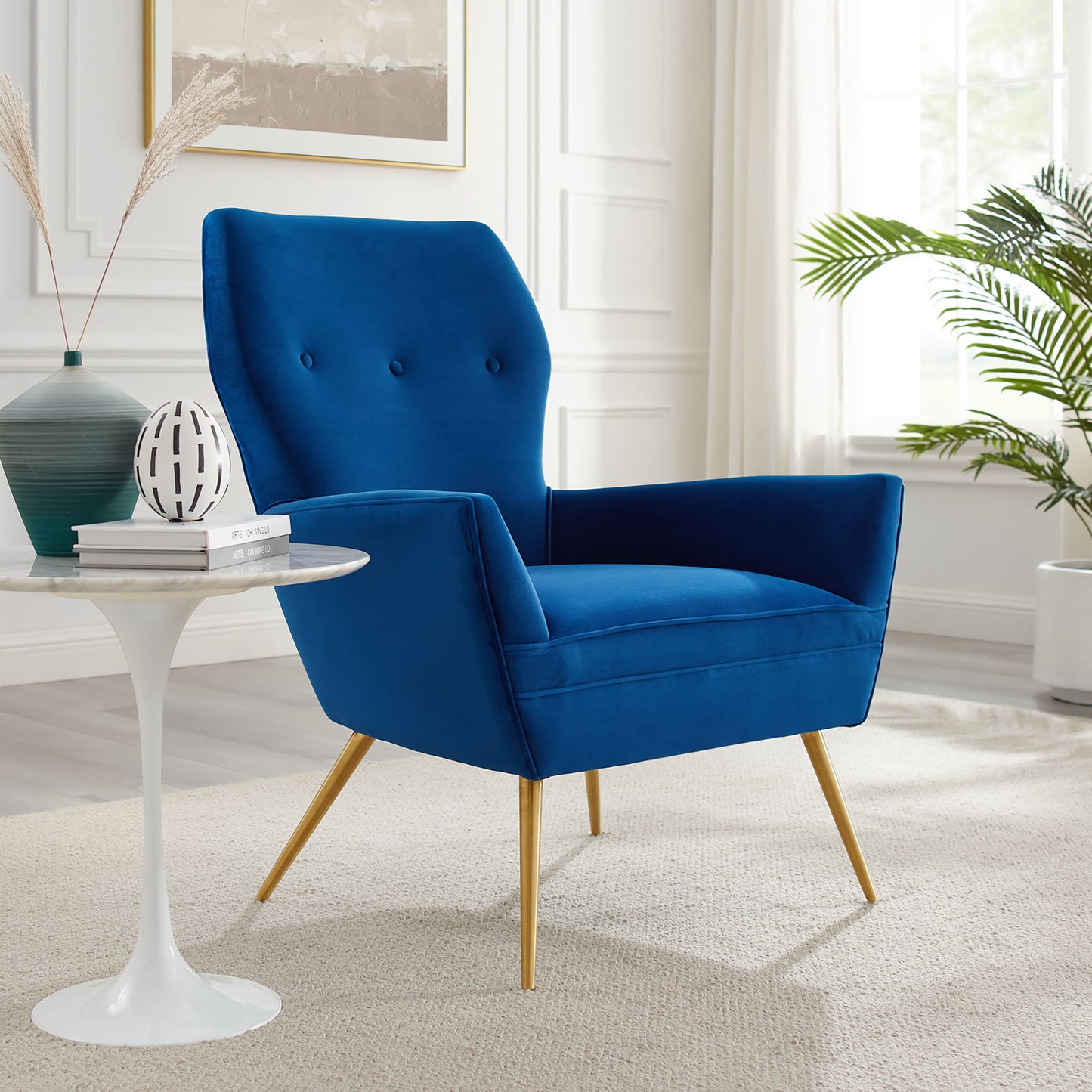 Navy velvet armchair with button-tufted back and gold tapered legs in a bright living room.