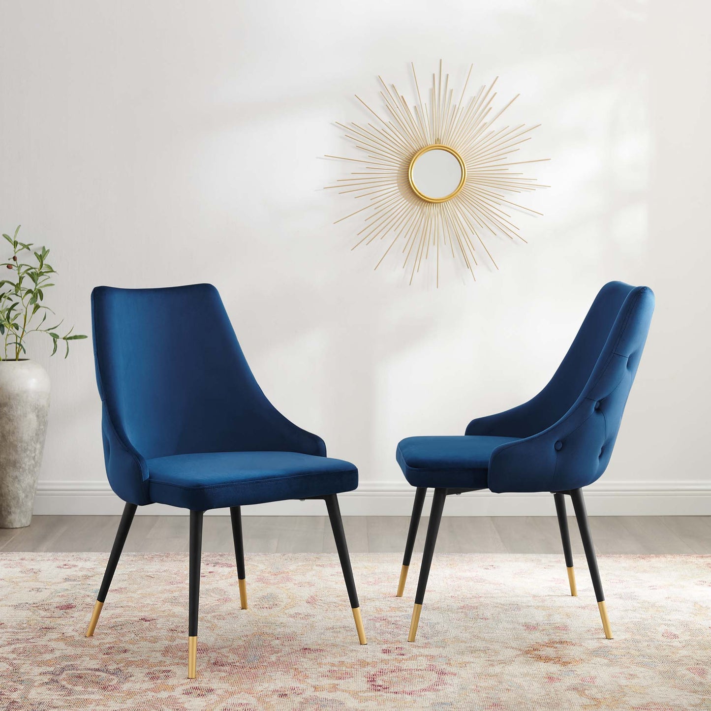 Two navy velvet dining side chairs with curved backs, black legs and gold tips, on a patterned rug under a sunburst mirror.