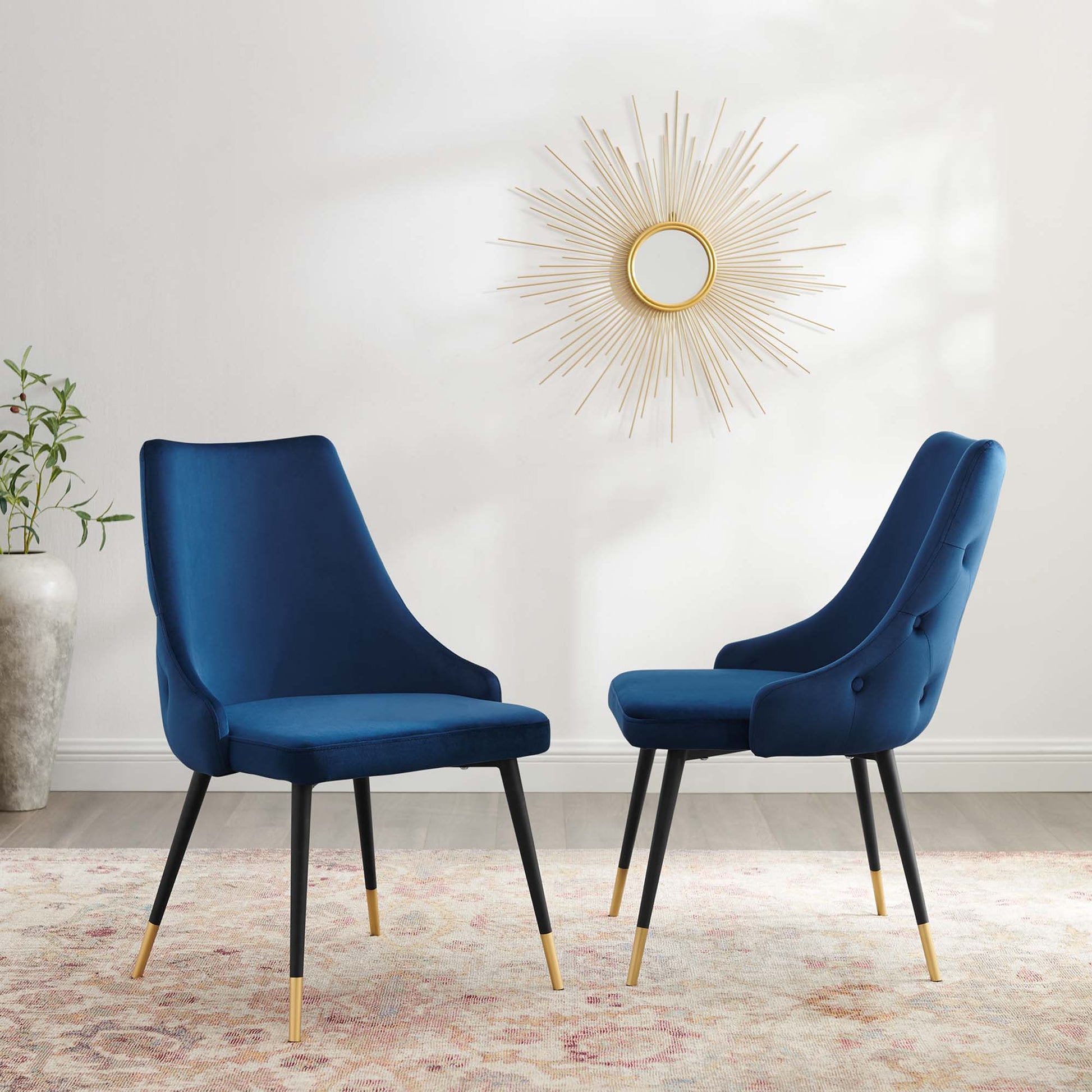 Two navy velvet dining side chairs with curved backs, black legs and gold tips, on a patterned rug under a sunburst mirror.