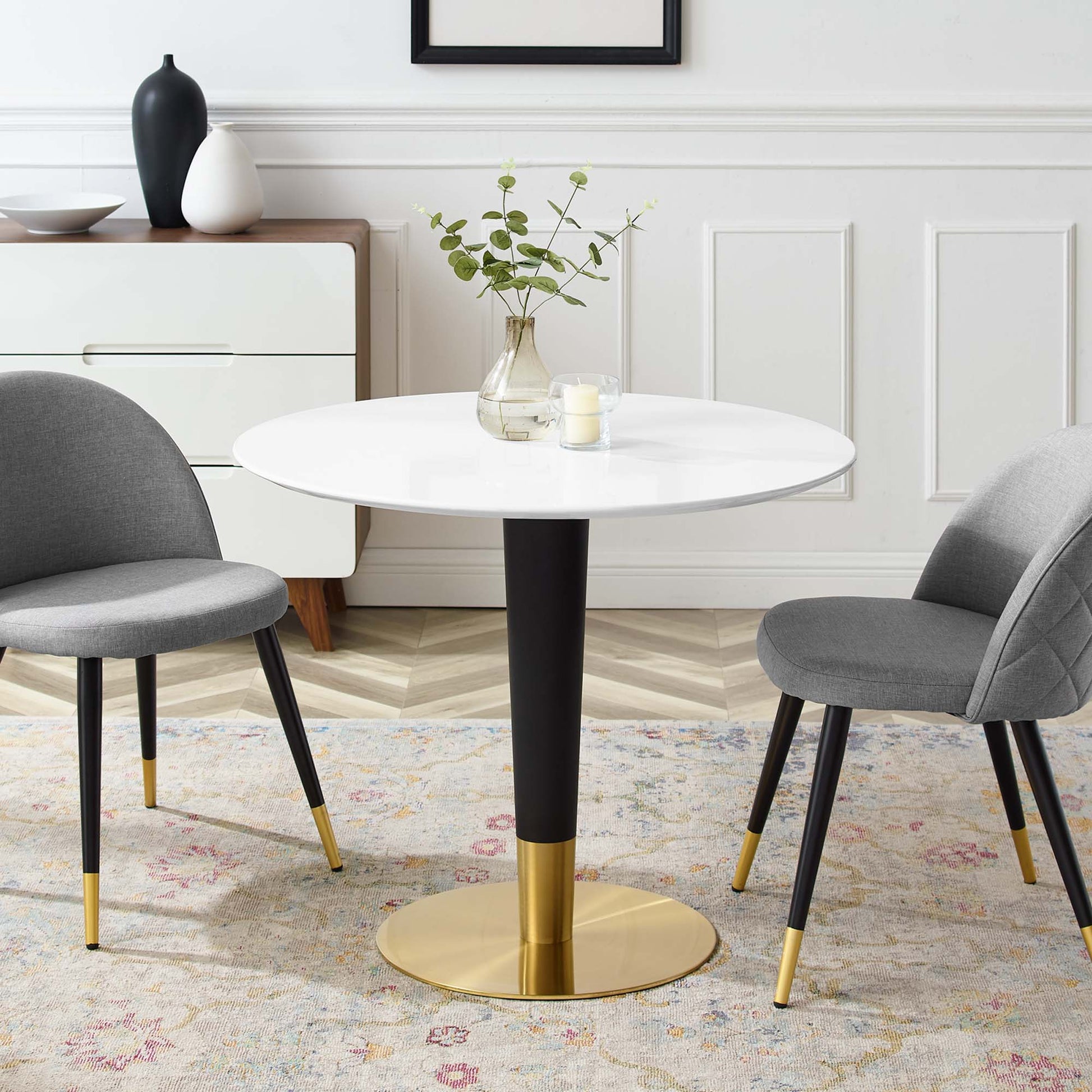 Round white dining table with black stem and gold pedestal base; gray upholstered chairs with gold-tipped legs on a pastel rug.