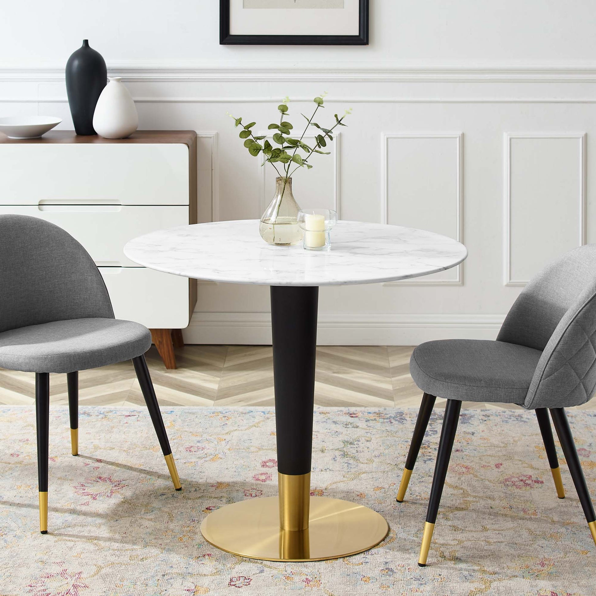 Round white marble dining table on black stem with gold pedestal, paired with grey upholstered chairs with gold-tipped legs.