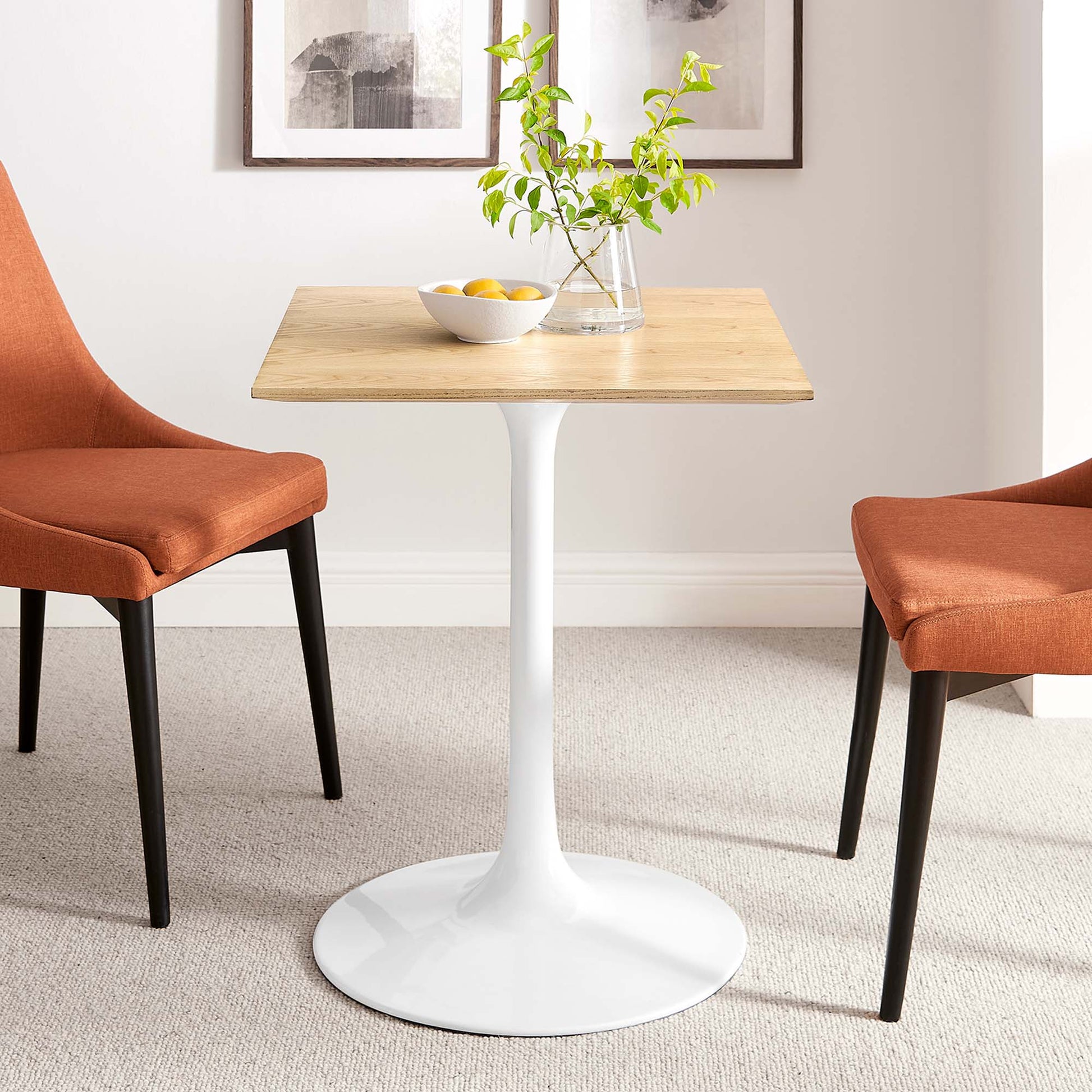 Wood-top pedestal dining table with white tulip base between orange upholstered chairs; plant vase and lemon bowl on tabletop.