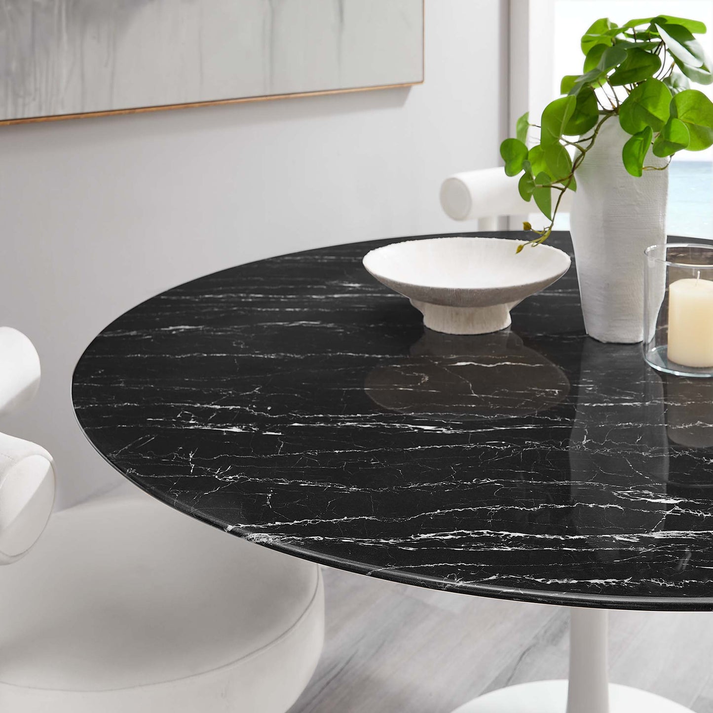 Round black faux marble-top dining table with white pedestal base, plant, bowl and candle decor.