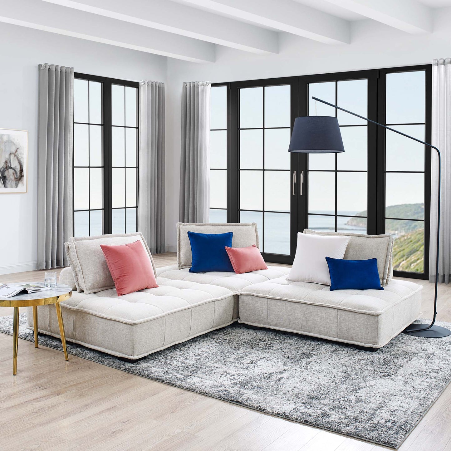 Beige modular sectional sofa with pink, blue, and white cushions in a bright living room with gray rug, black-framed windows, and an arc lamp.