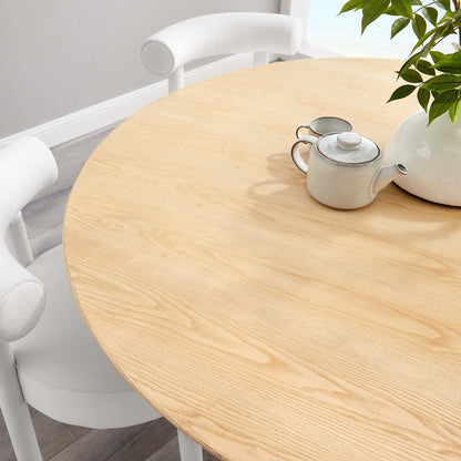 Round natural wood dining table with teapot, cup, and leafy plant centerpiece.