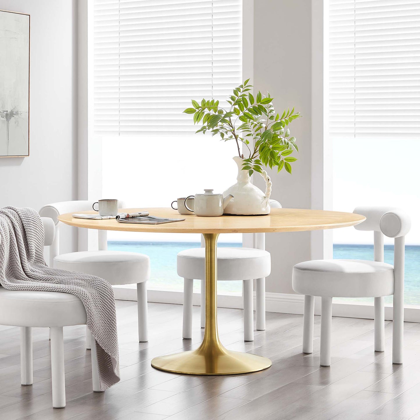 Round natural wood dining table with gold pedestal base in a bright sunlit dining nook, white stools, plant centerpiece.