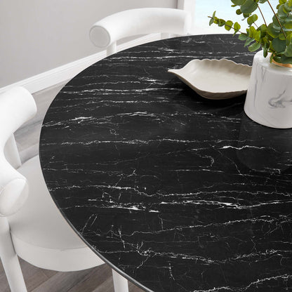 Round black faux marble dining table with white veining, white modern chairs, beige shell-shaped dish & potted plant.