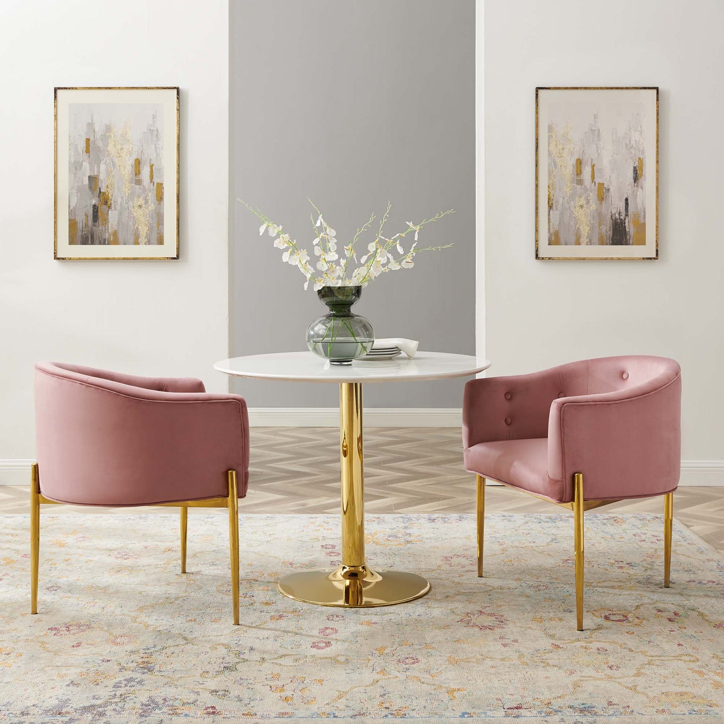 Pink velvet accent chairs with gold legs sit around a round white table with a gold pedestal base, vase of white flowers.