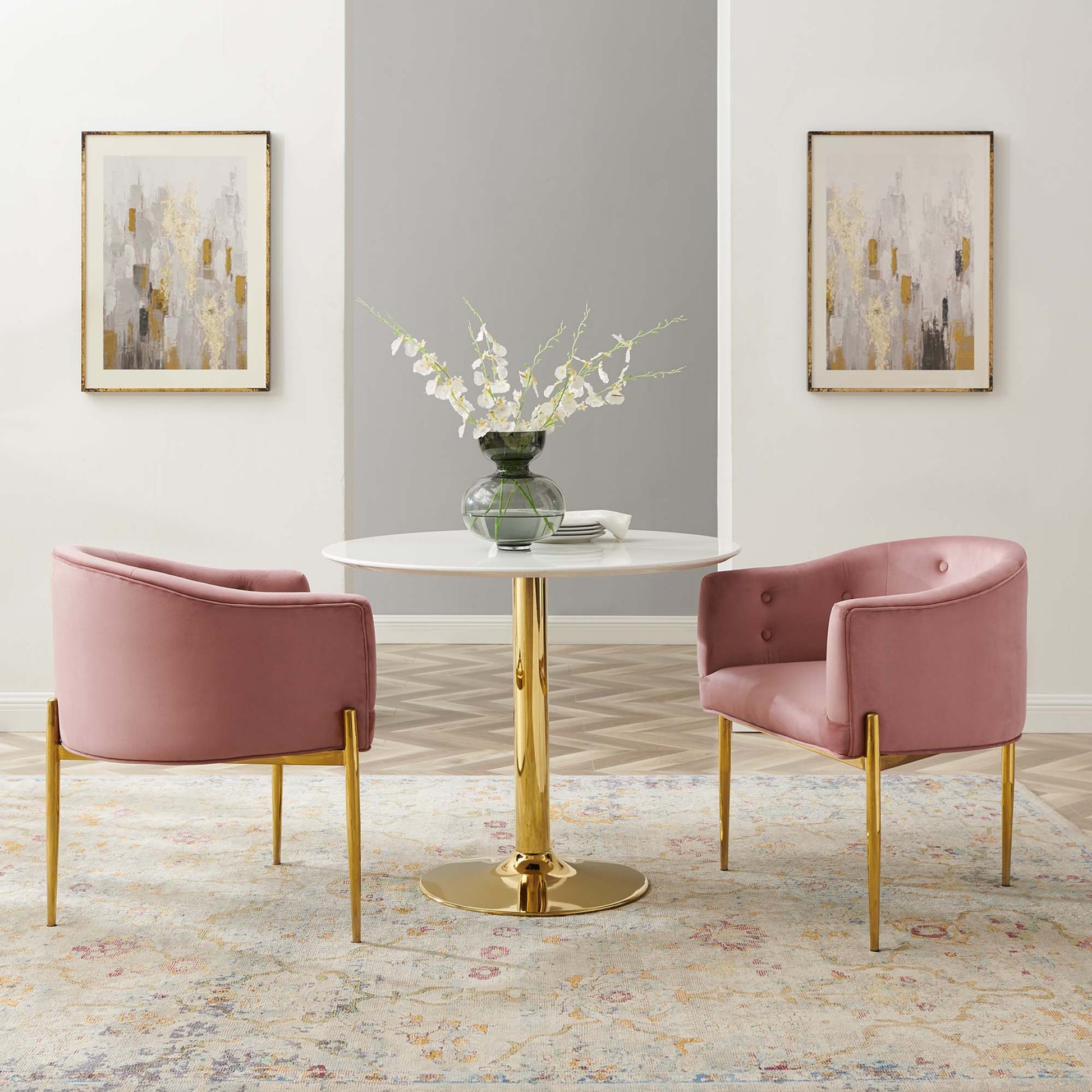 Pink velvet accent chairs with gold legs sit around a round white table with a gold pedestal base, vase of white flowers.