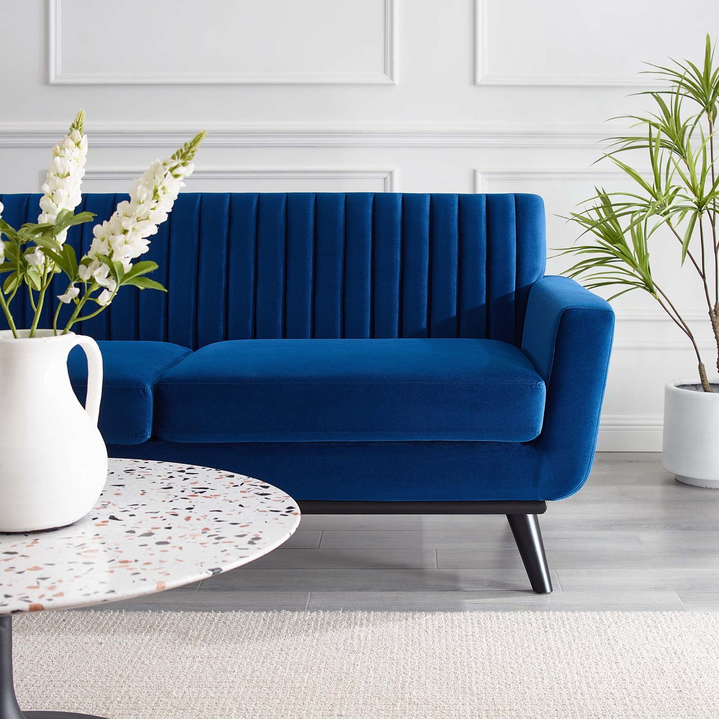 Blue velvet sofa & vertical tufting & black angled legs in a bright white-walled living room, terrazzo table & white vase.