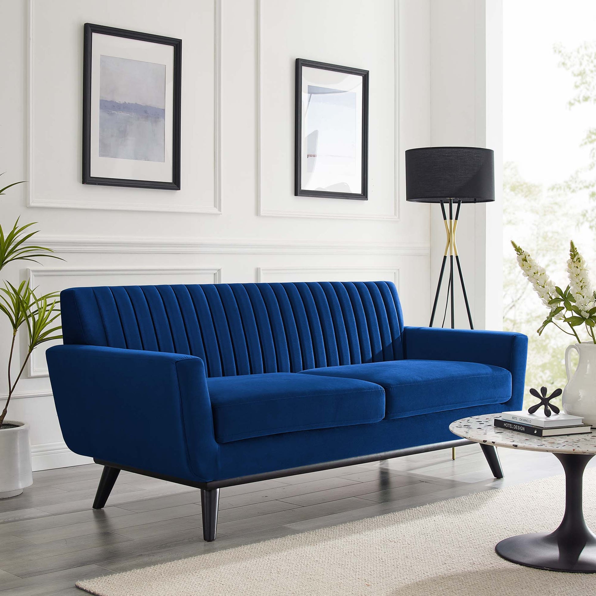 Navy blue velvet loveseat with vertical channel tufting and mid-century legs in a bright living room with abstract art.