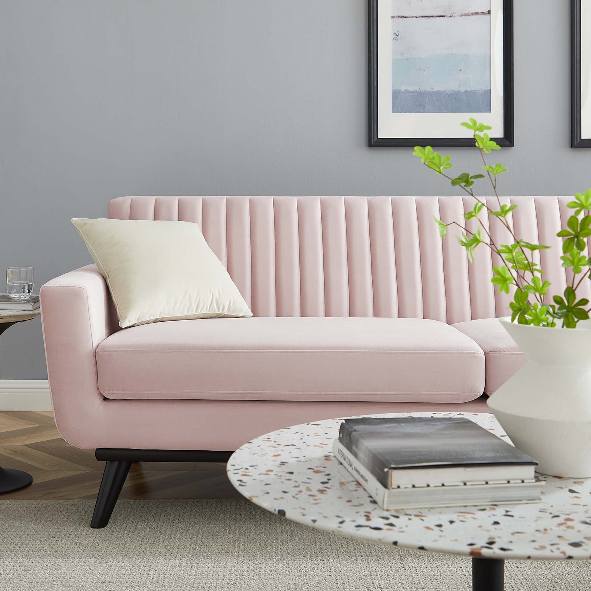 Pastel pink velvet sofa with vertical channels, black legs, in modern living room with terrazzo coffee table and greenery.