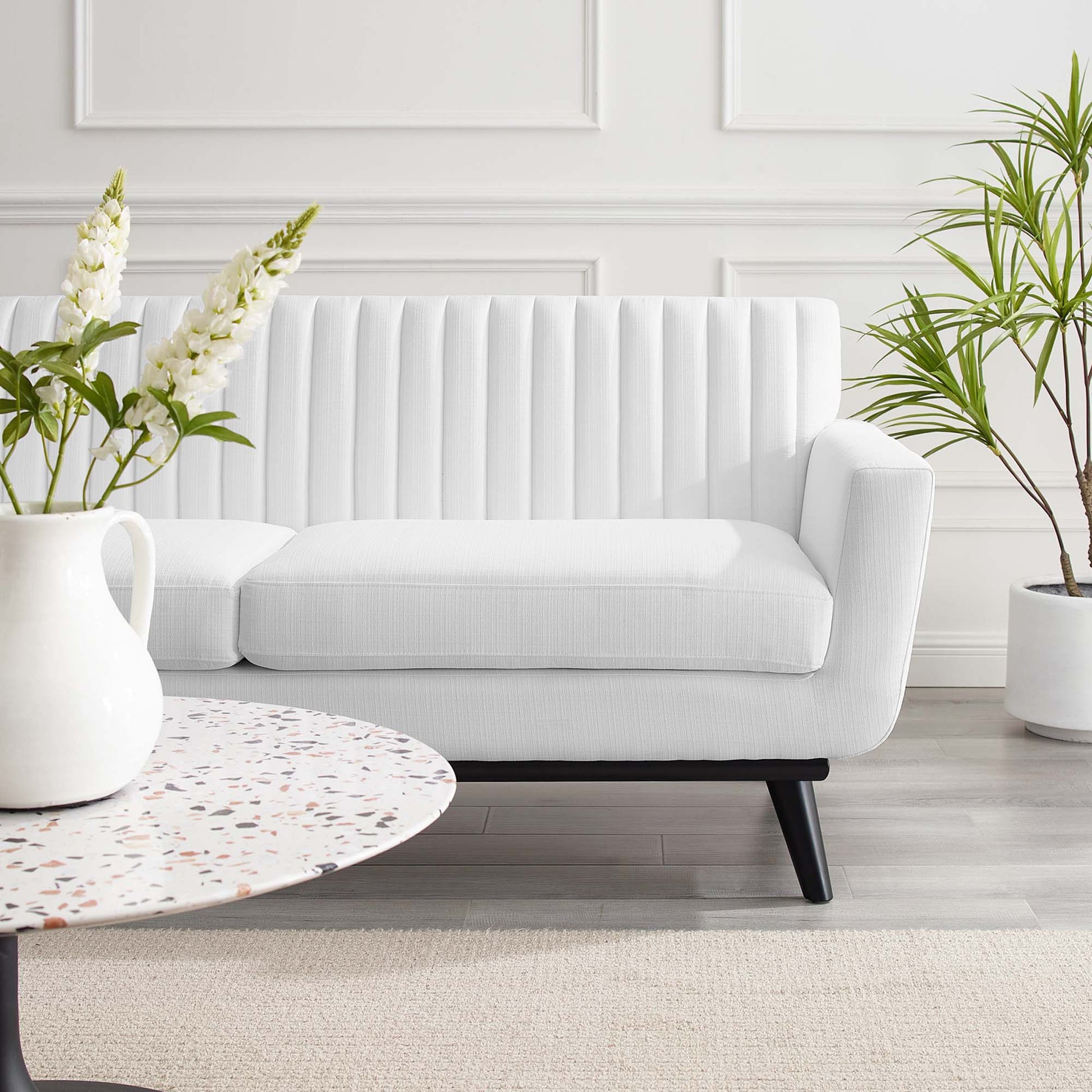 White mid-century loveseat with vertical channeling and black legs in a bright living room with a round terrazzo coffee table.