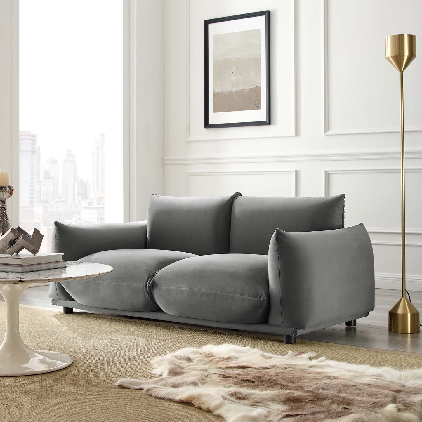 Grey velvet loveseat with thick cushions in a modern living room, with a gold floor lamp, fur rug, and circular coffee table.