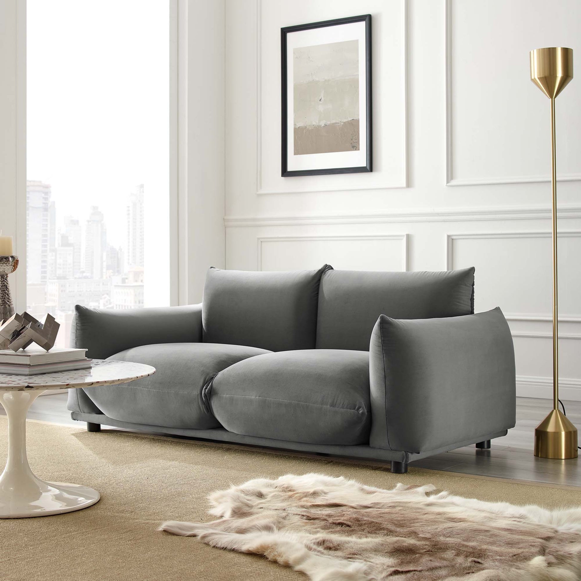 Grey velvet loveseat with thick cushions in a modern living room, with a gold floor lamp, fur rug, and circular coffee table.