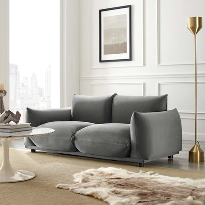 Grey velvet loveseat with thick cushions in a modern living room, with a gold floor lamp, fur rug, and circular coffee table.