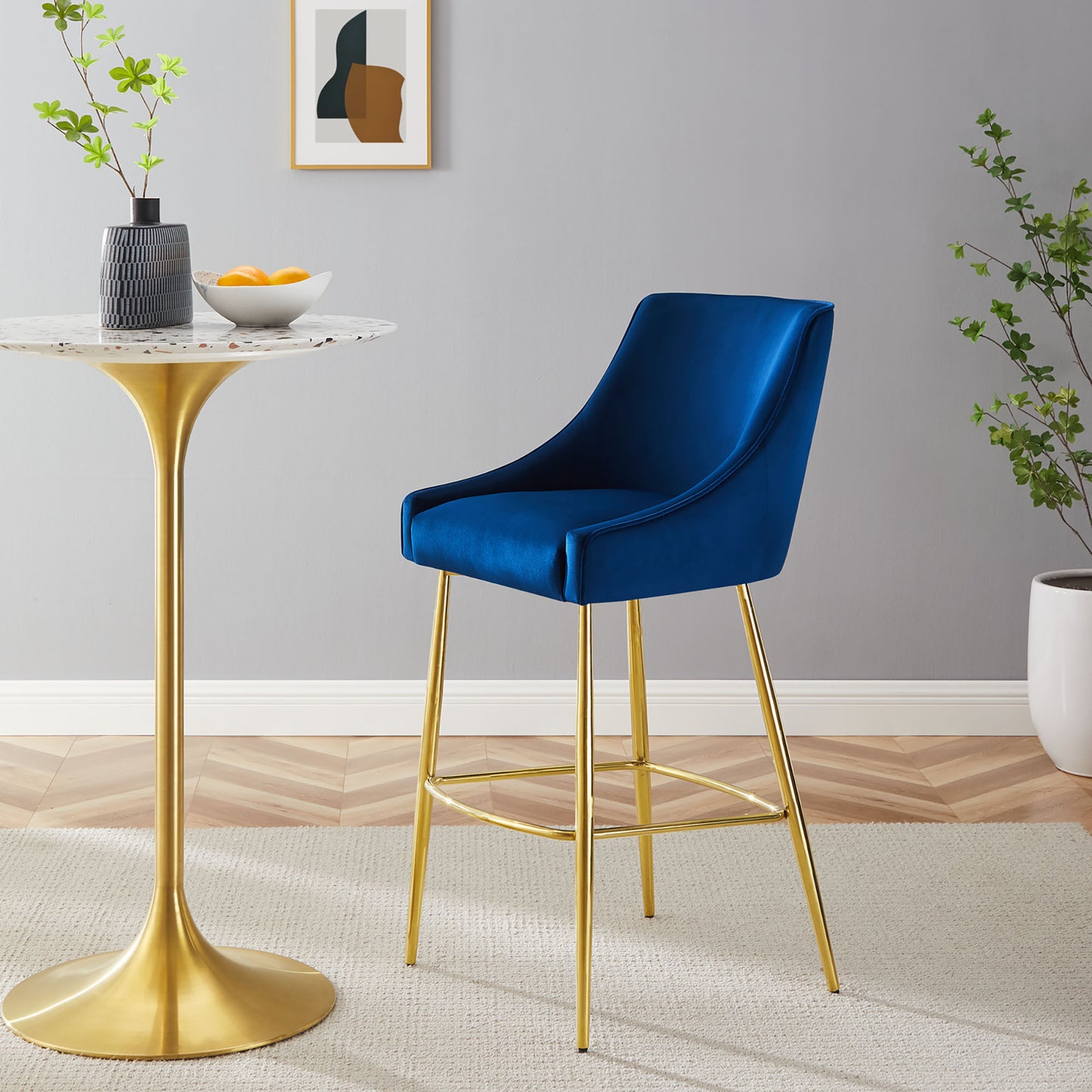 Navy velvet bar stool with gold legs and a curved back beside a marble-top pedestal table in a modern dining nook.