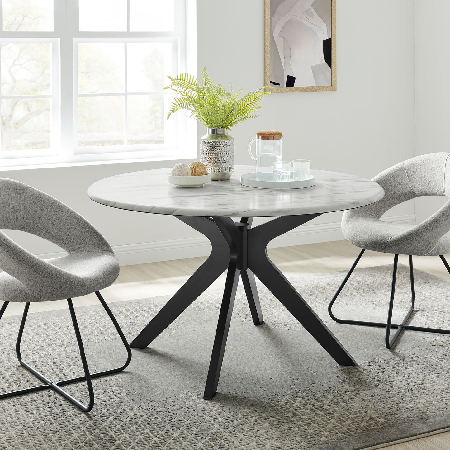 Round marble-top dining table with a black angular pedestal base, paired with gray upholstered chairs and a fern centerpiece.