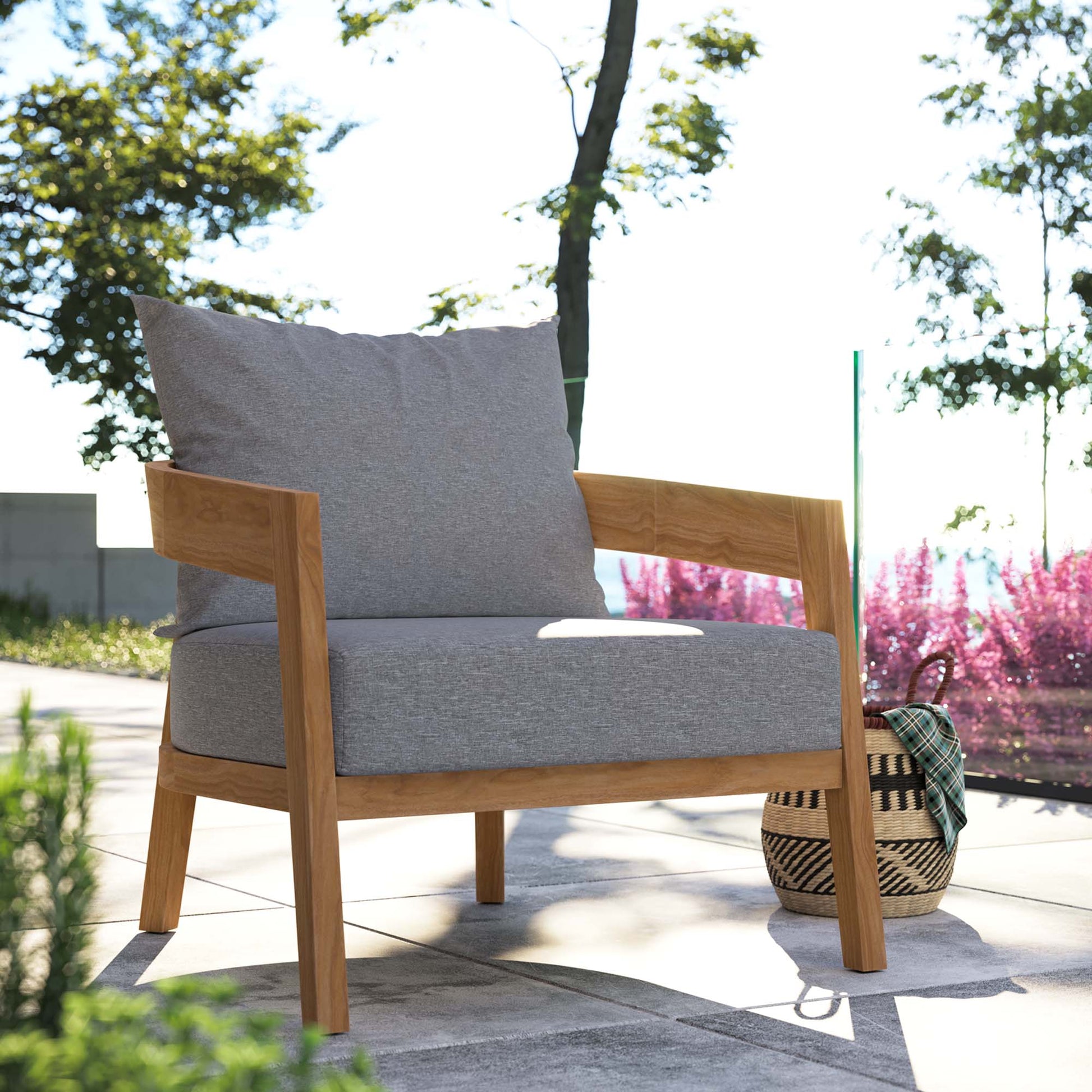 Outdoor teak wood patio armchair with light gray cushions on a sunlit patio, with a woven basket beside.