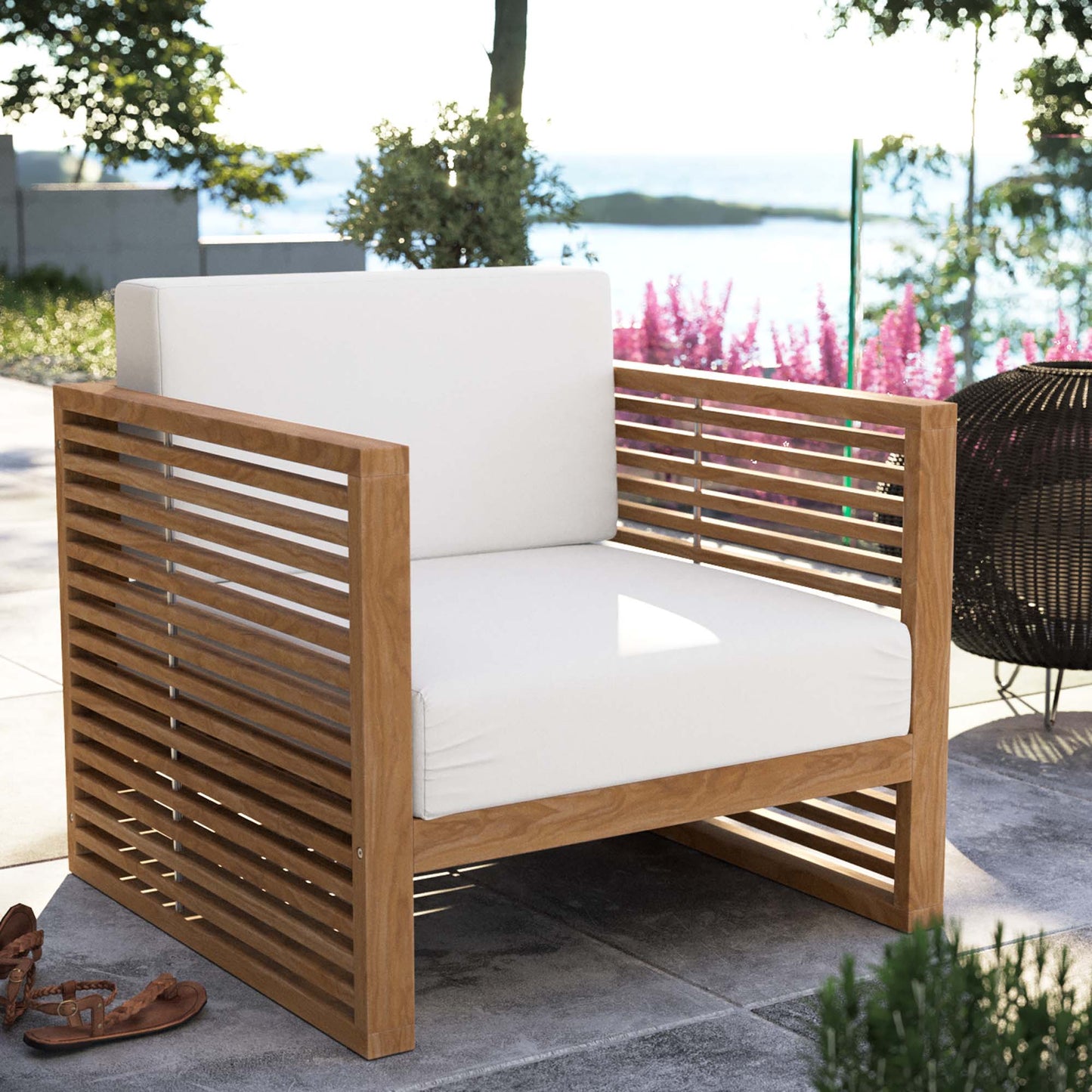 Teak wood outdoor armchair with white cushions, slatted sides and back, on a sunny patio by the water.
