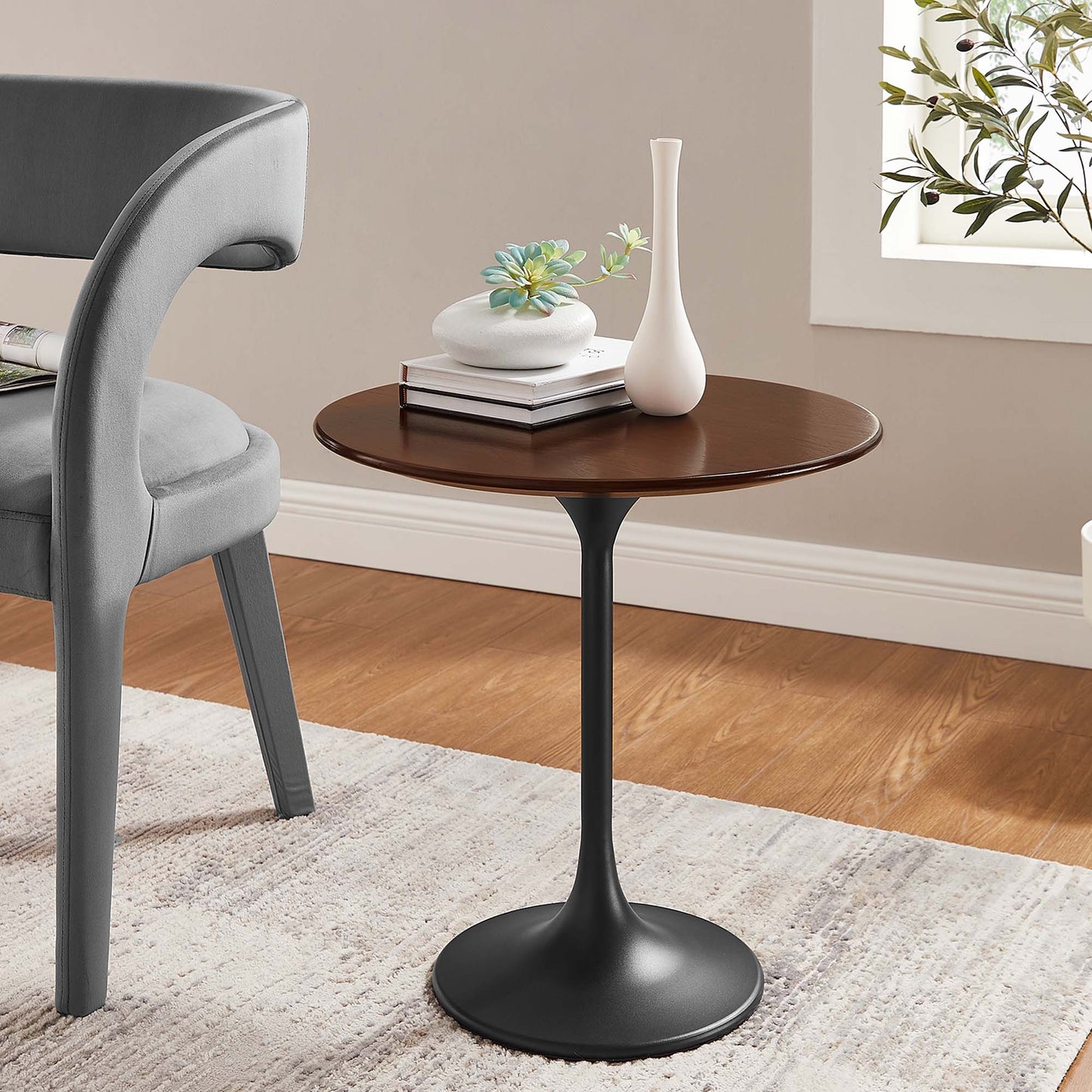 Round walnut-brown wood top side table on a black tulip base; gray upholstered chair beside, with vase and books.