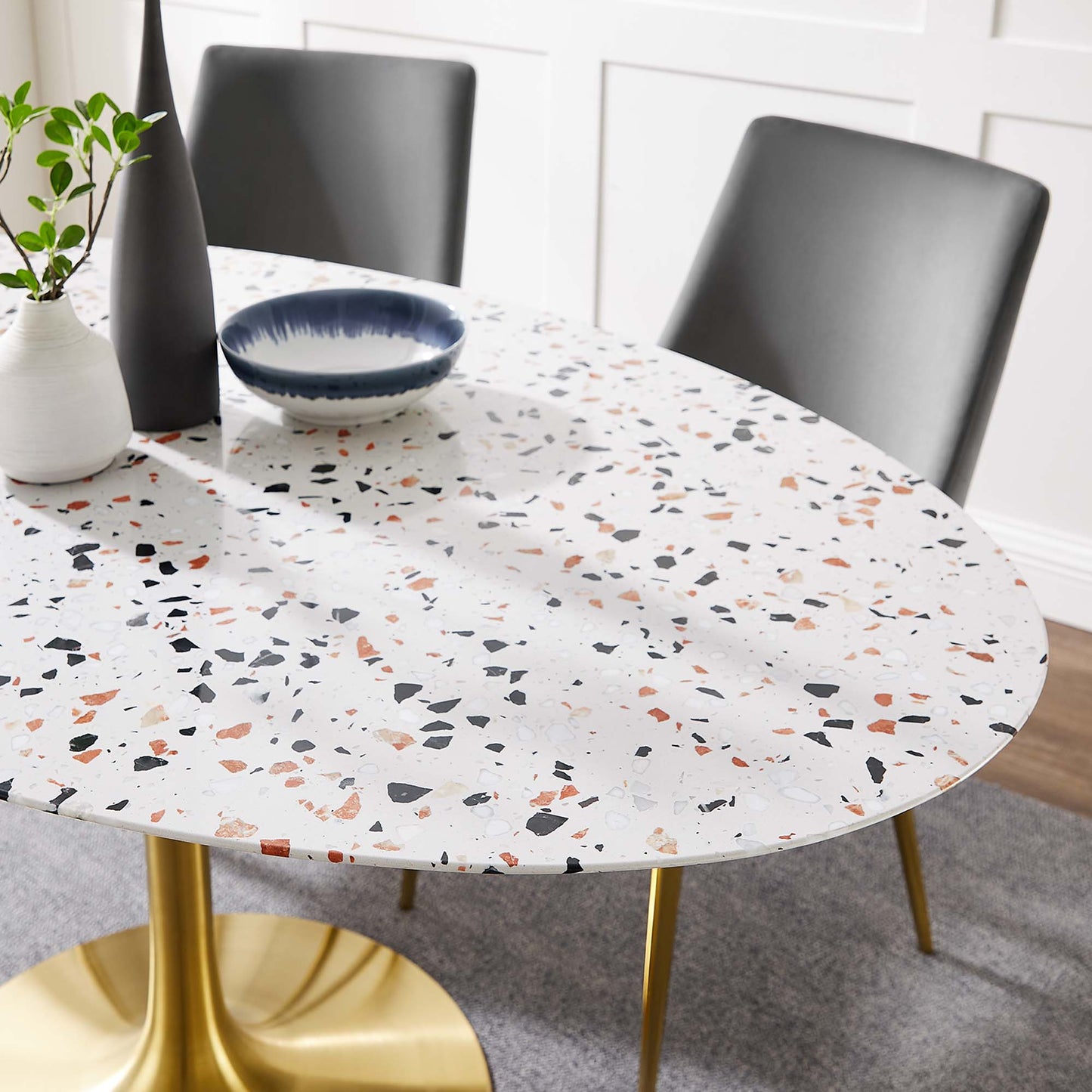 Oval terrazzo dining table with gold legs, blue bowl, black vase, white vase with greenery, gray upholstered chairs.