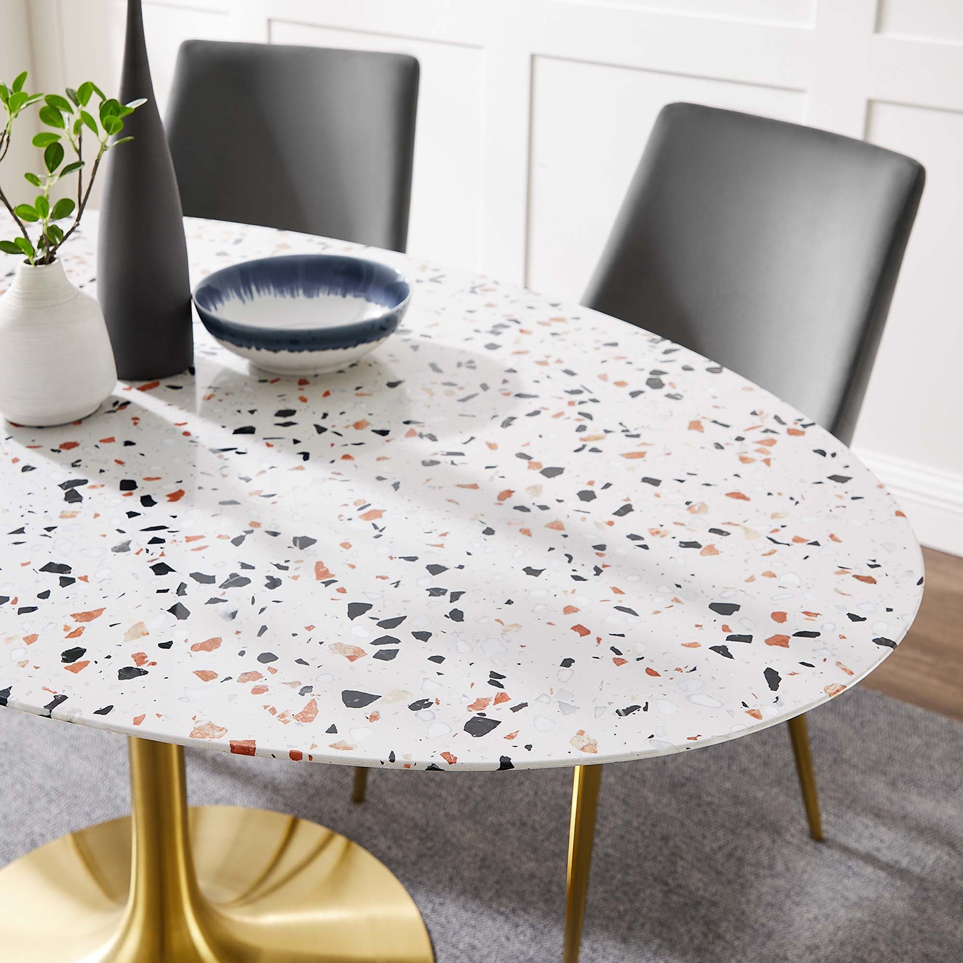 Oval terrazzo dining table with gold legs, blue bowl, black vase, white vase with greenery, gray upholstered chairs.