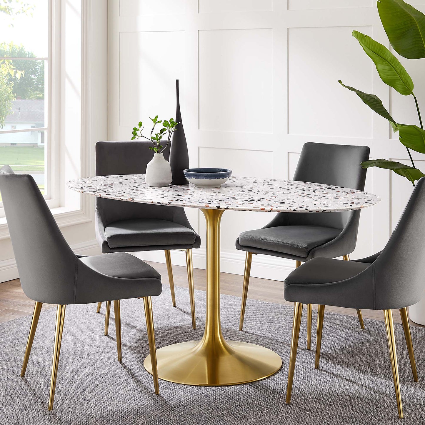 Oval terrazzo dining table with a white speckled top on a gold pedestal base, surrounded by gray upholstered chairs in a bright dining room.