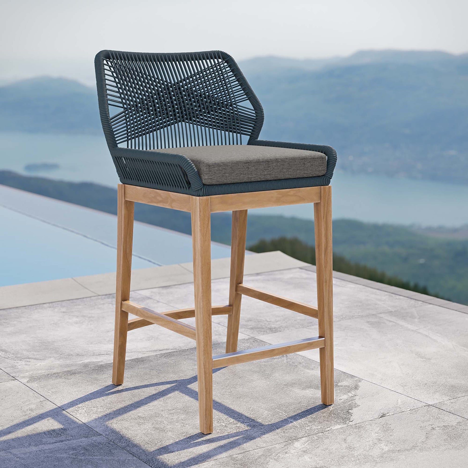 Outdoor bar stool with gray woven back and seat, gray cushion, teak wood legs on a lakeside patio.