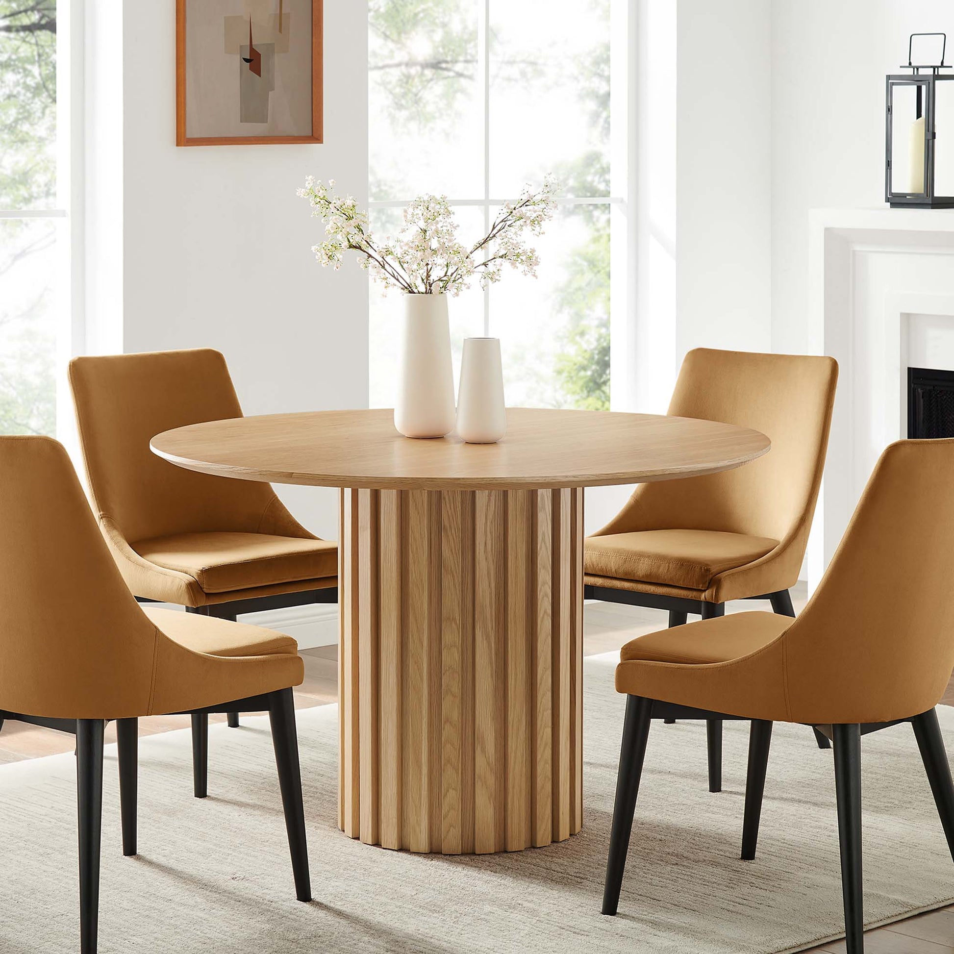 Oak round dining table with vertical slat pedestal, surrounded by tan upholstered chairs in a bright modern dining set.