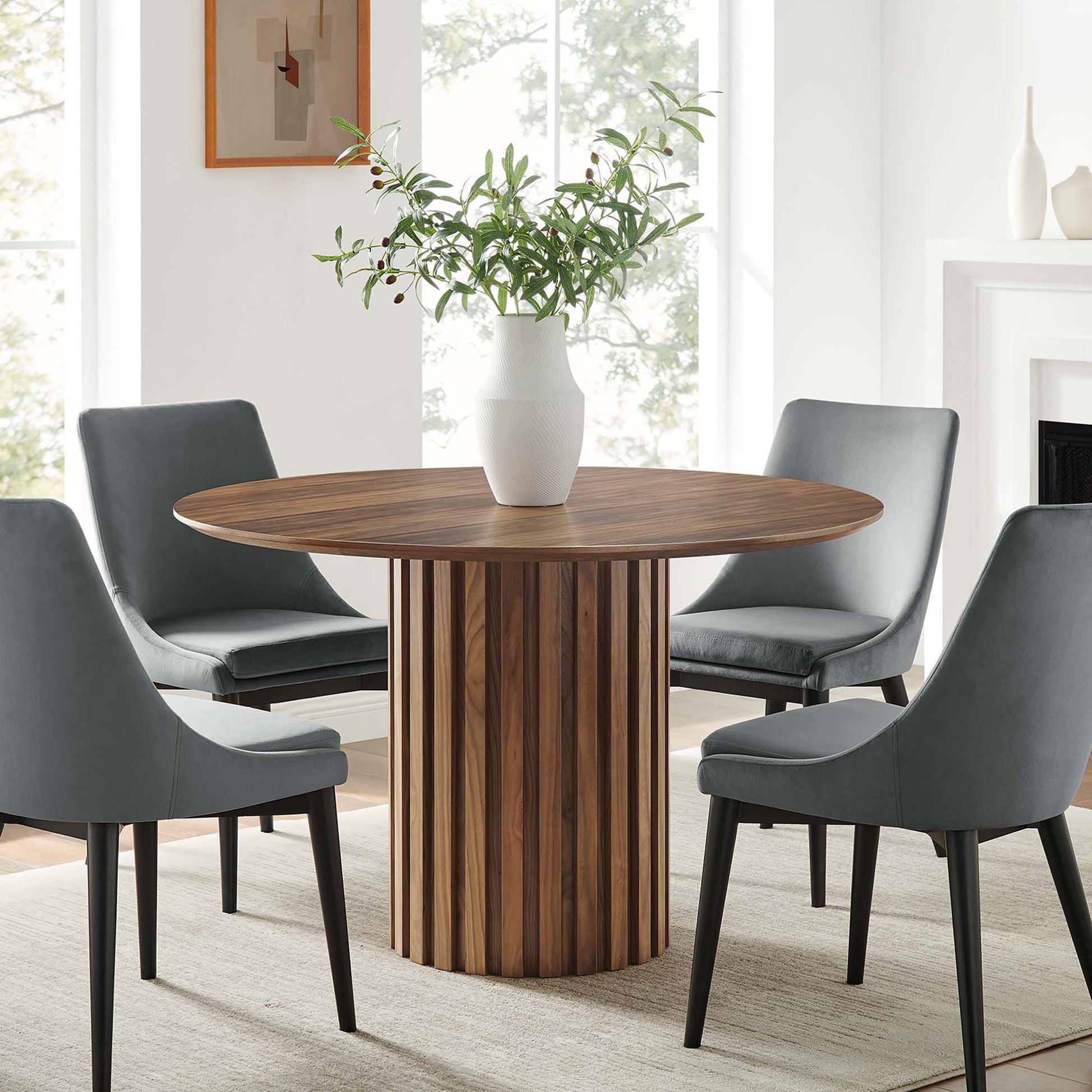 Round walnut dining table with vertical slat pedestal, gray upholstered chairs, and a white vase centerpiece.