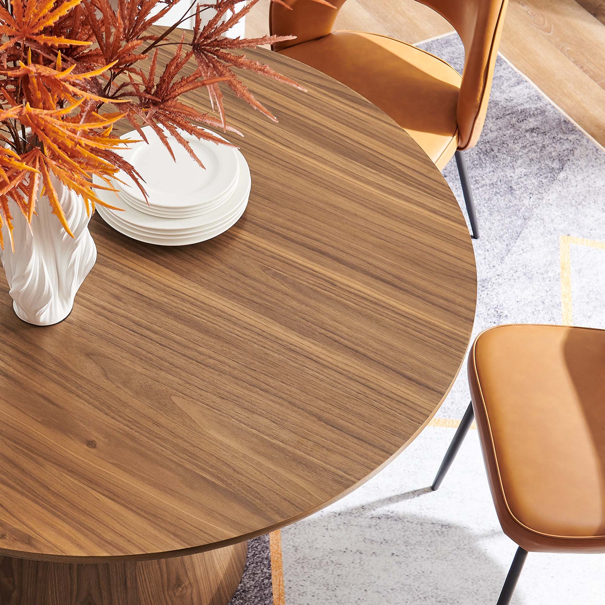 Round walnut wood dining table with white vase & orange foliage, stacked white plates, surrounded by tan leather chairs.
