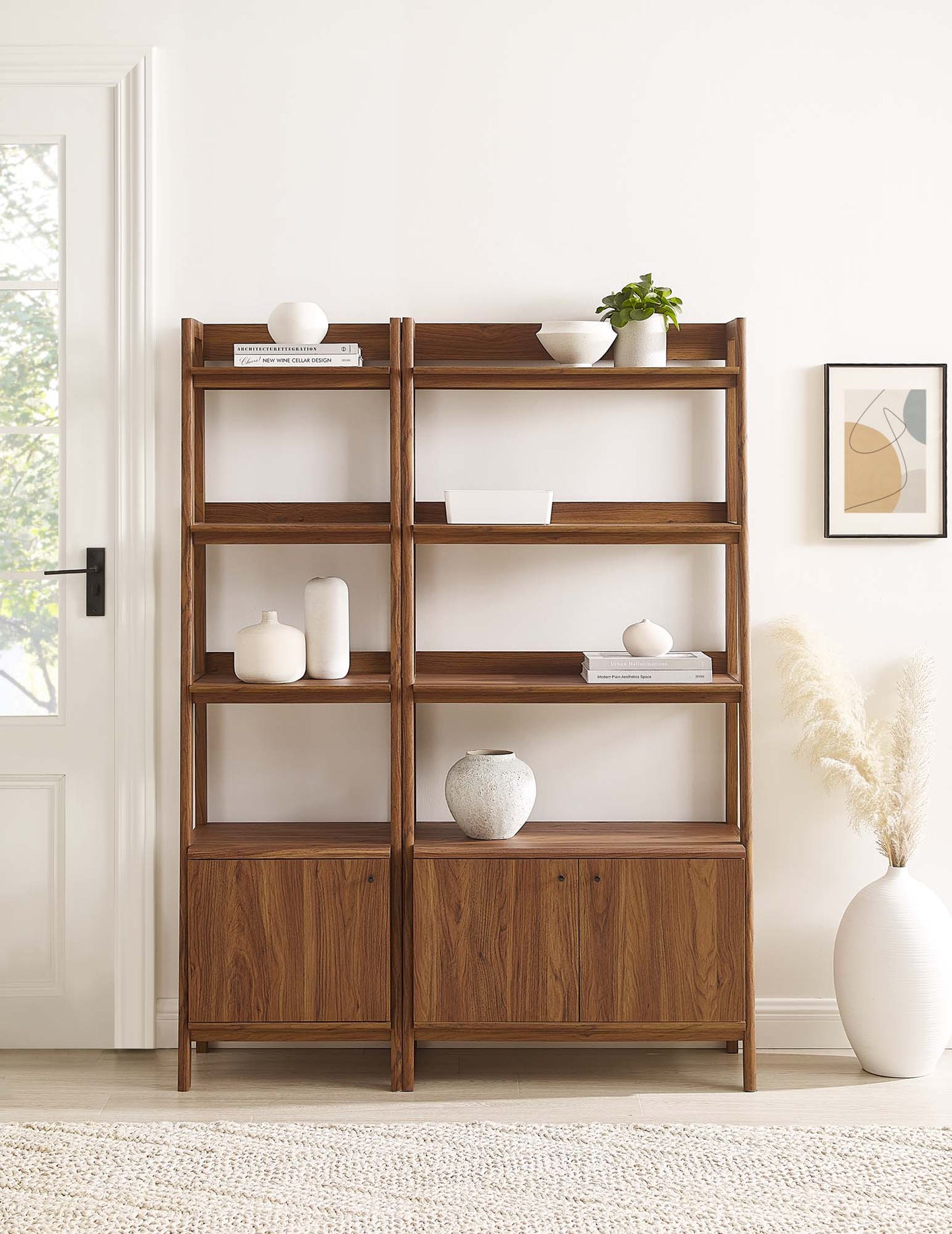 Walnut wood pair of bookcases with four open shelves above a cabinet base, decorated with white ceramics, books, and a plant.