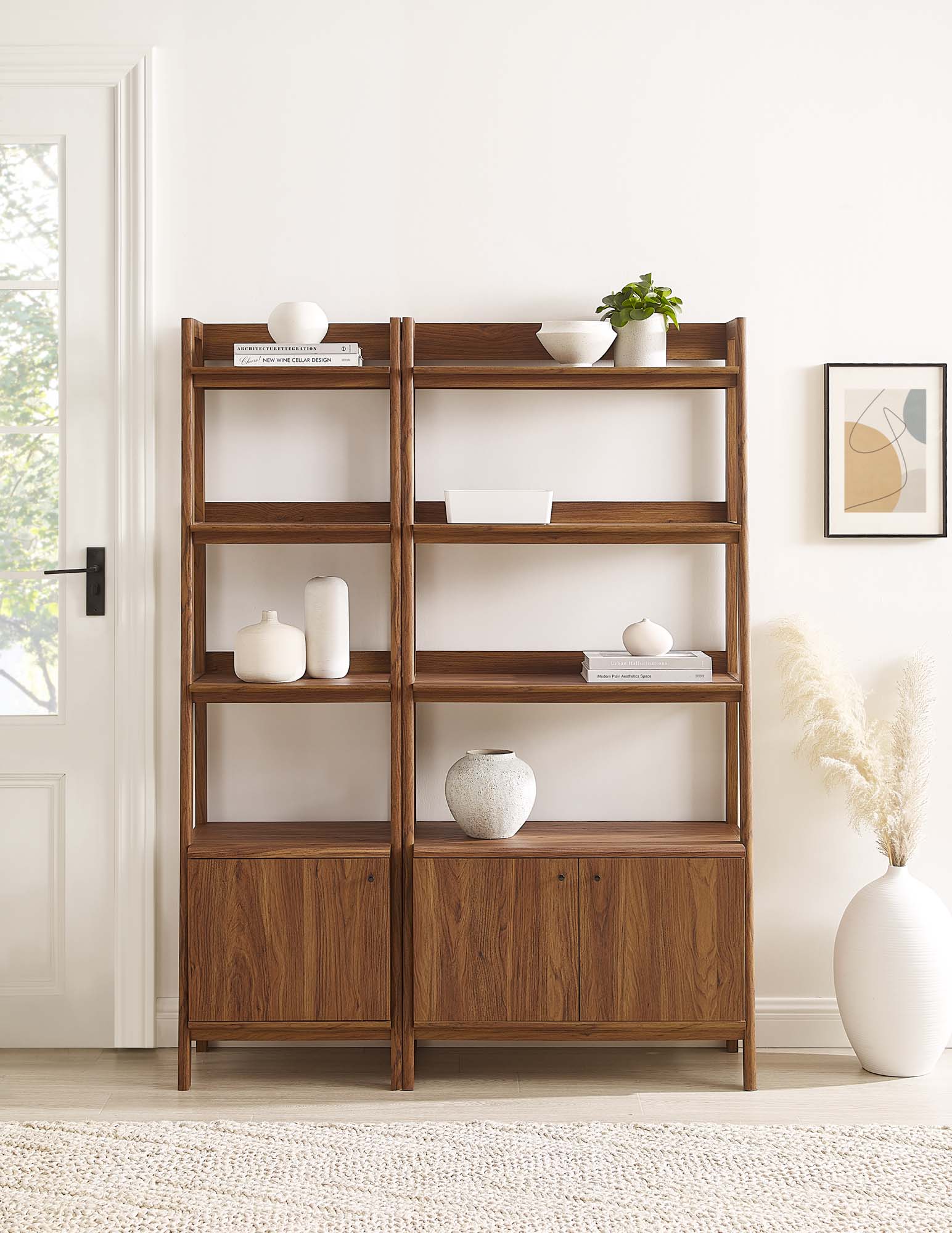 Walnut wood pair of bookcases with four open shelves above a cabinet base, decorated with white ceramics, books, and a plant.