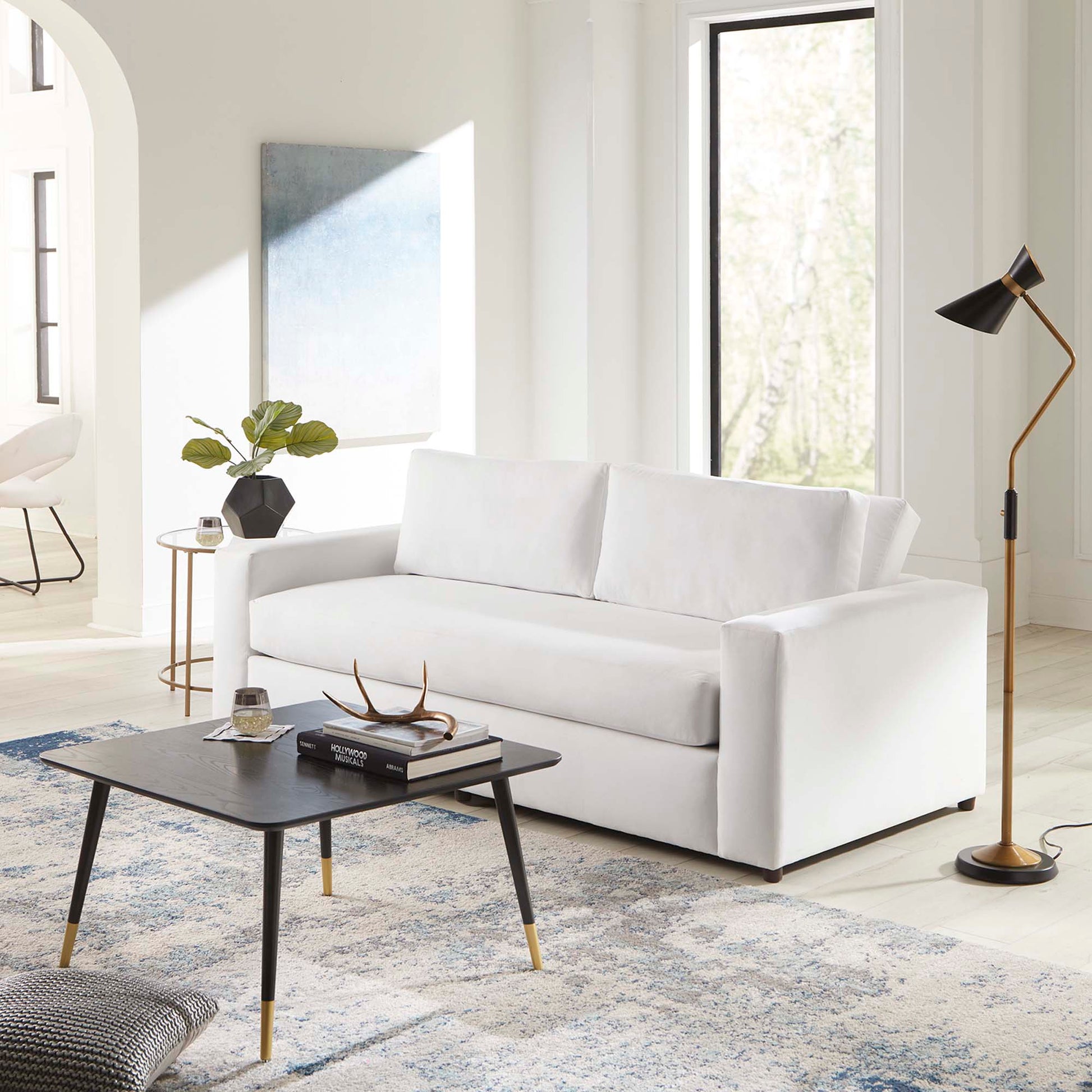 Modern white velvet sofa with clean lines in a bright living room, black coffee table with gold-tipped legs.