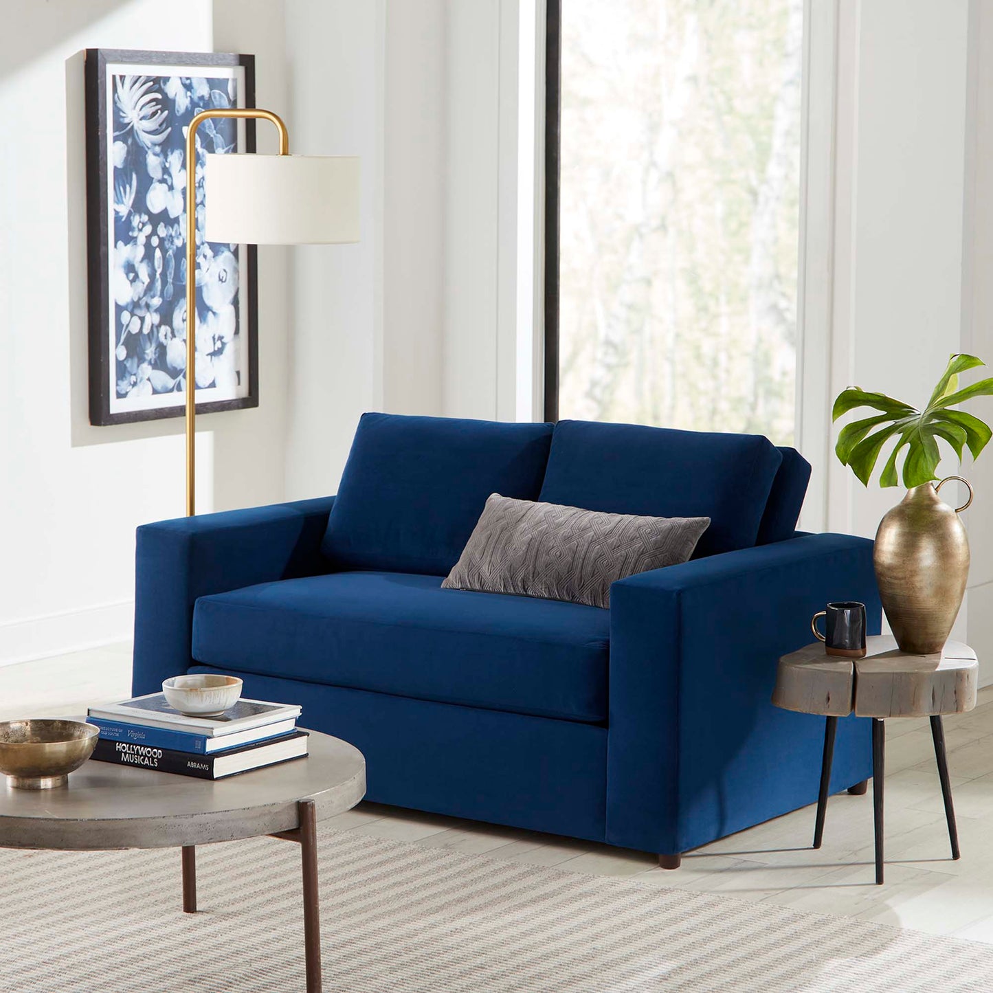 Deep navy velvet loveseat with a gray lumbar pillow in a bright living room; gold arc floor lamp, round coffee table, and side table.