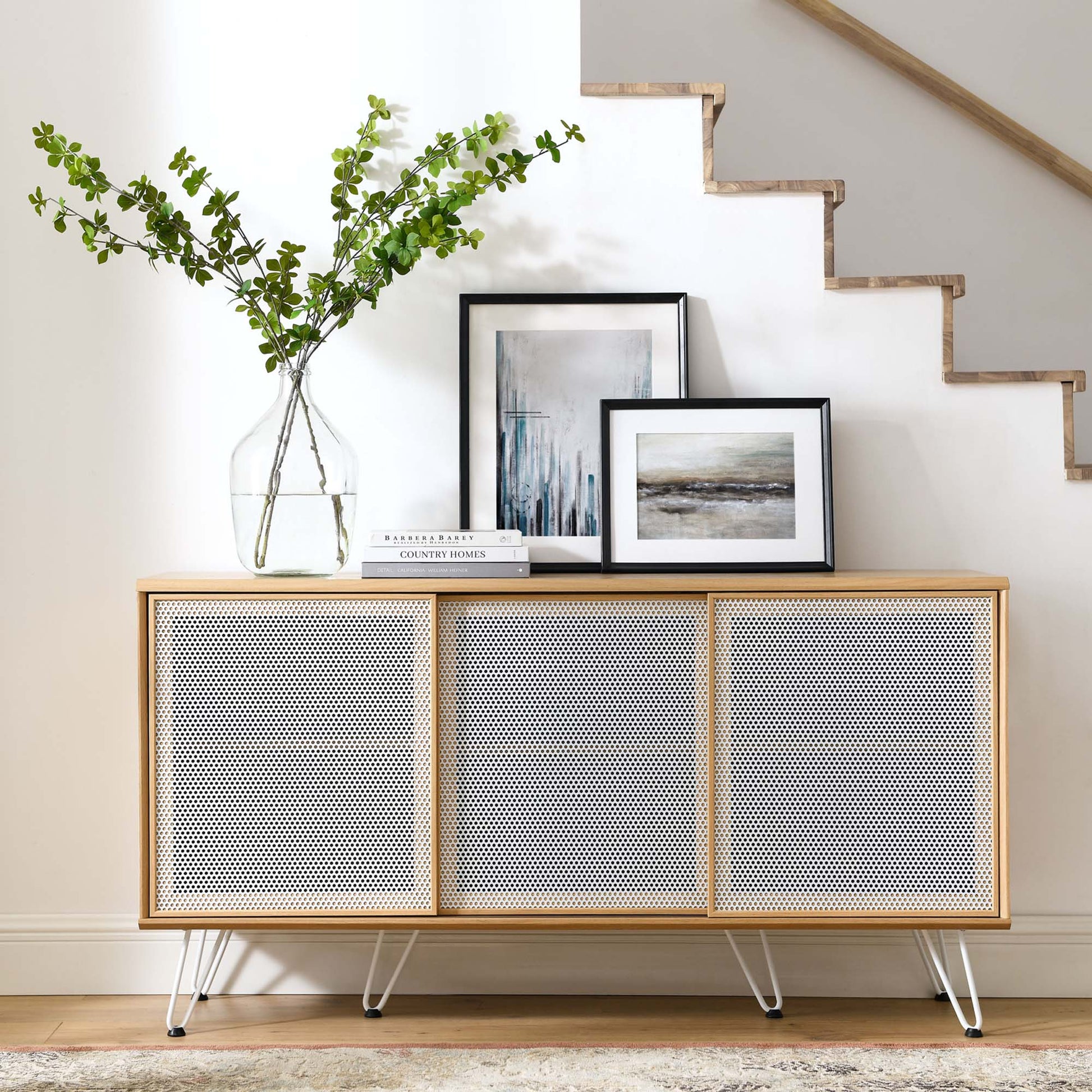 Oak-framed sideboard with four perforated panels, slim metal legs, topped with a vase of greenery and framed art.