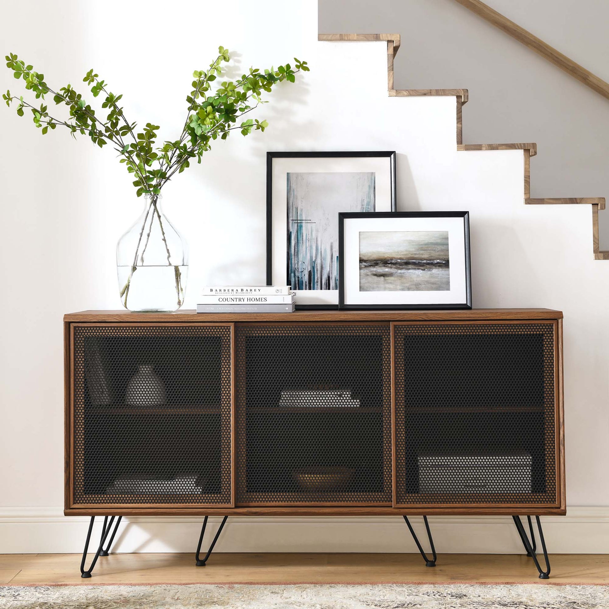 Walnut sideboard with three mesh-front cabinets on hairpin legs, topped with a vase of greenery and framed artwork.