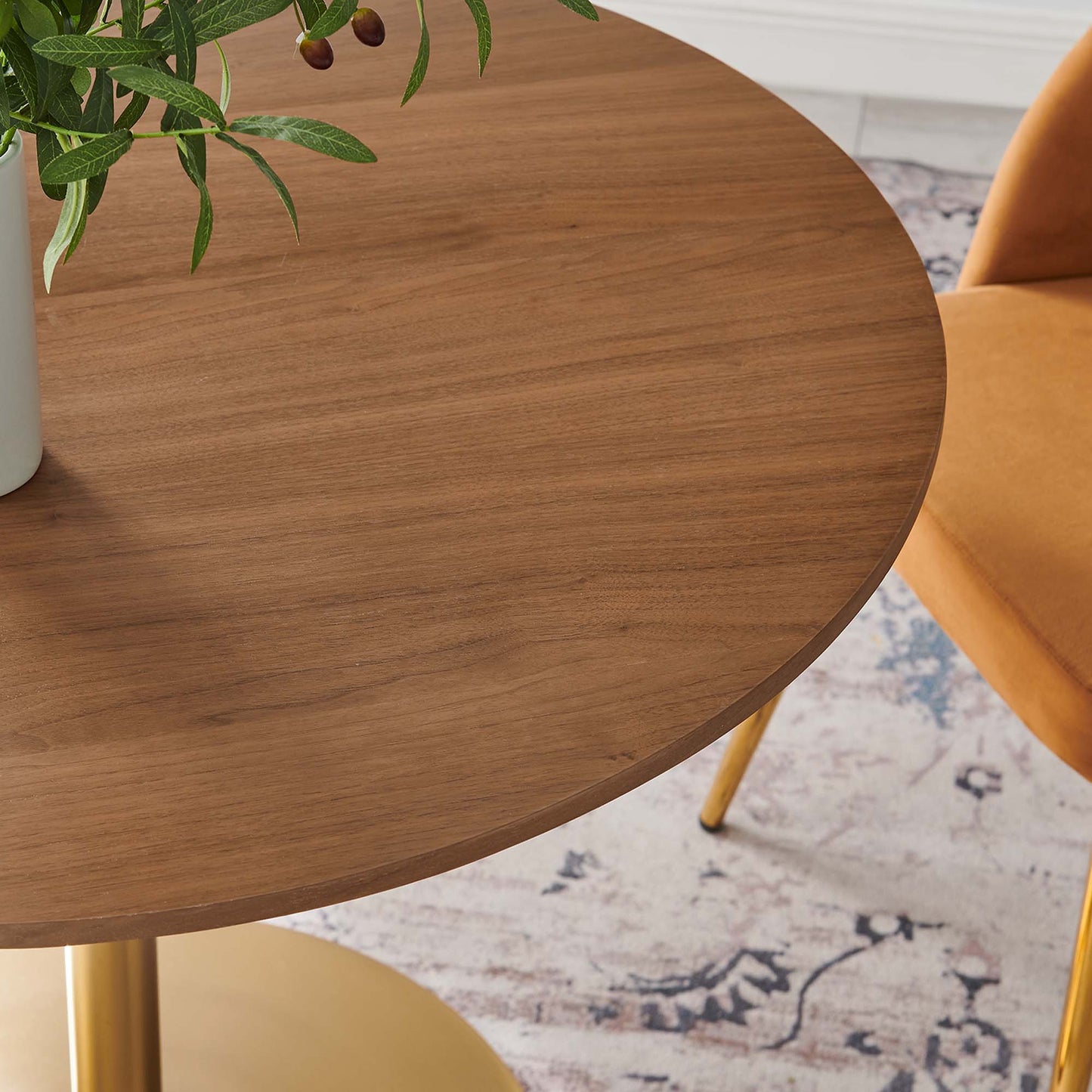 Round walnut wood table with gold metal legs, plant on top, and an orange chair in the background.