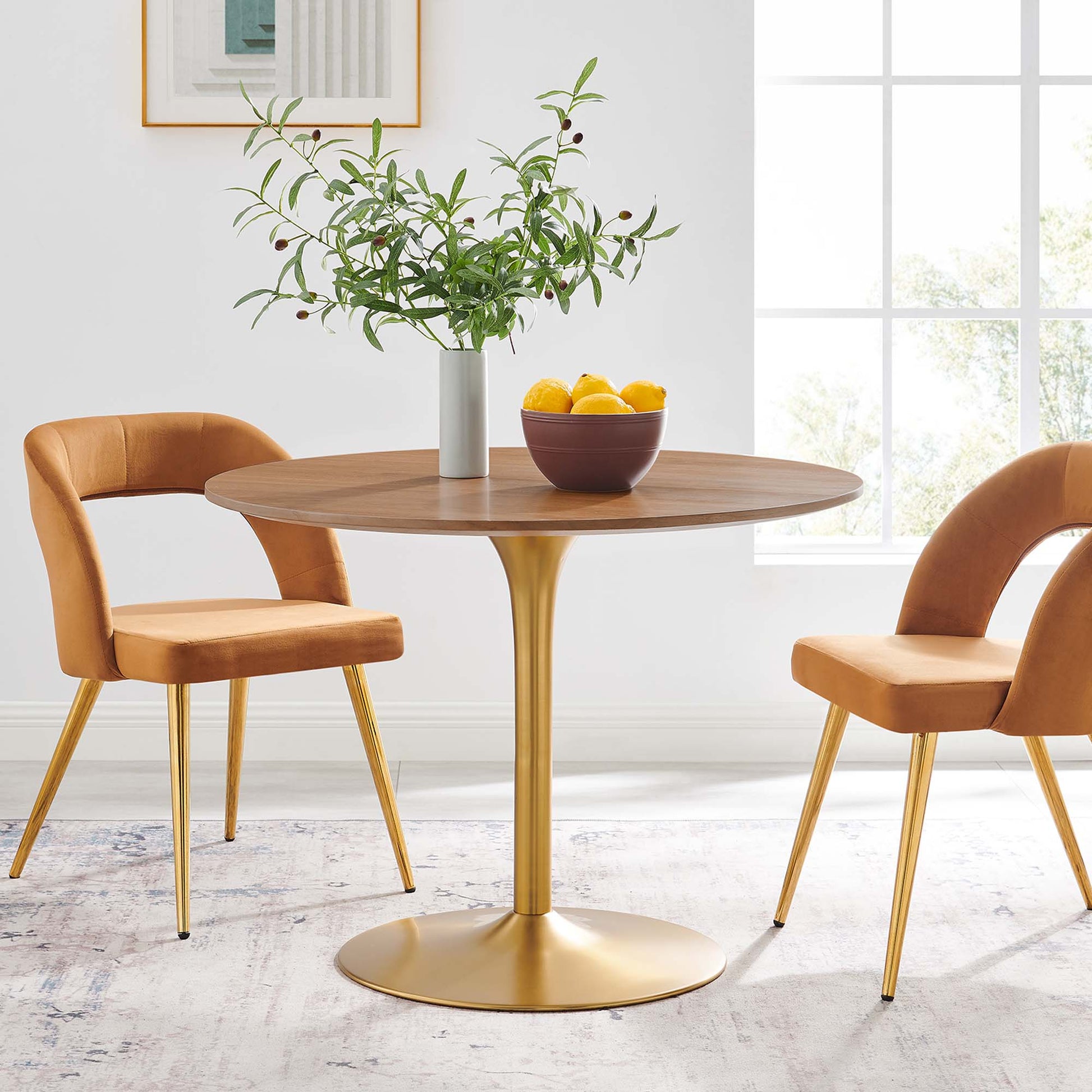 Round walnut dining table with gold pedestal base, tan upholstered mid-century chairs; greenery vase and orange bowl.