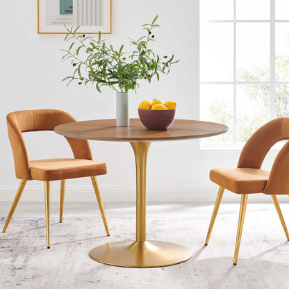 Round walnut dining table with gold pedestal base, tan upholstered mid-century chairs; greenery vase and orange bowl.