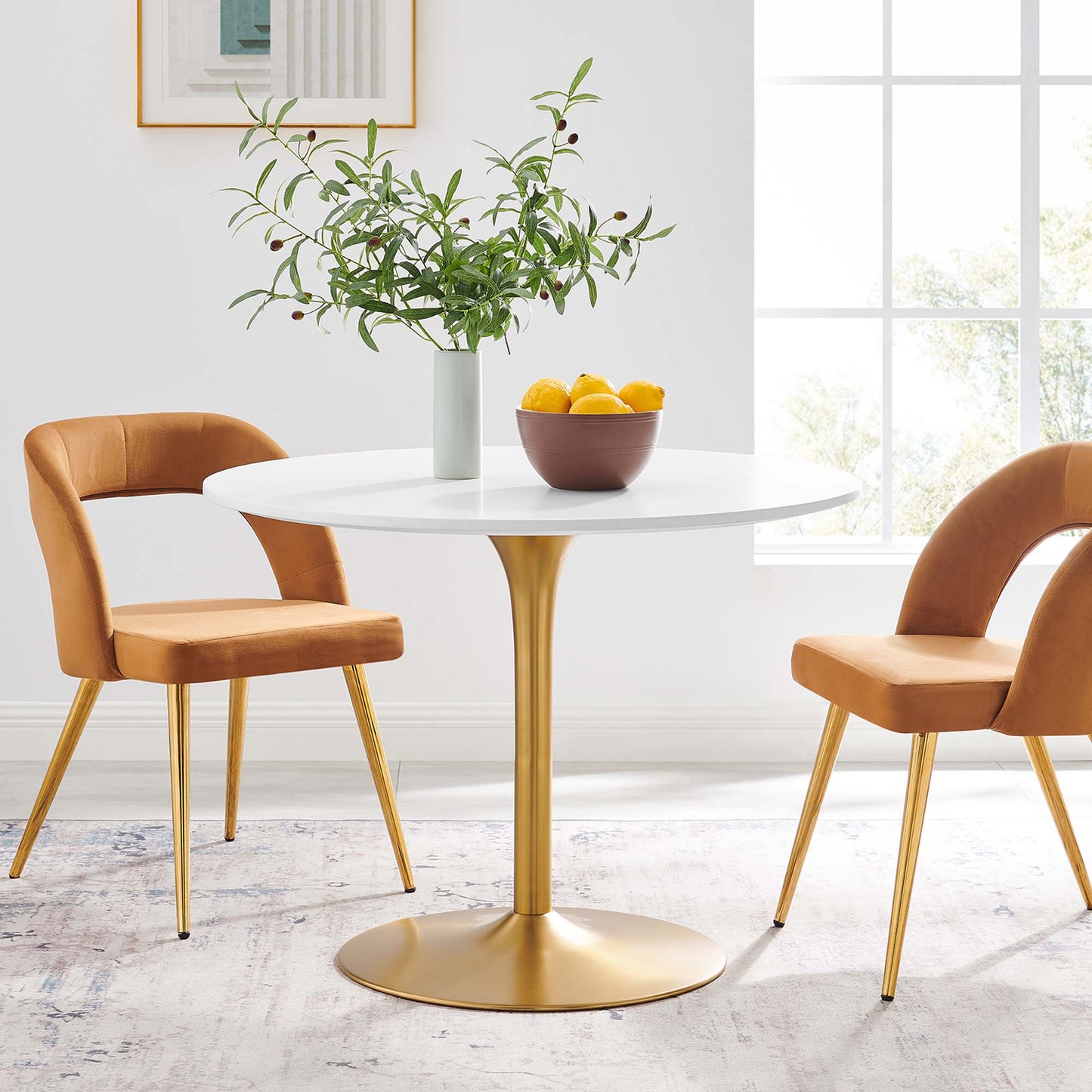 White round dining table with gold pedestal base, orange upholstered chairs, vase of greenery, lemons, sunlit dining nook.