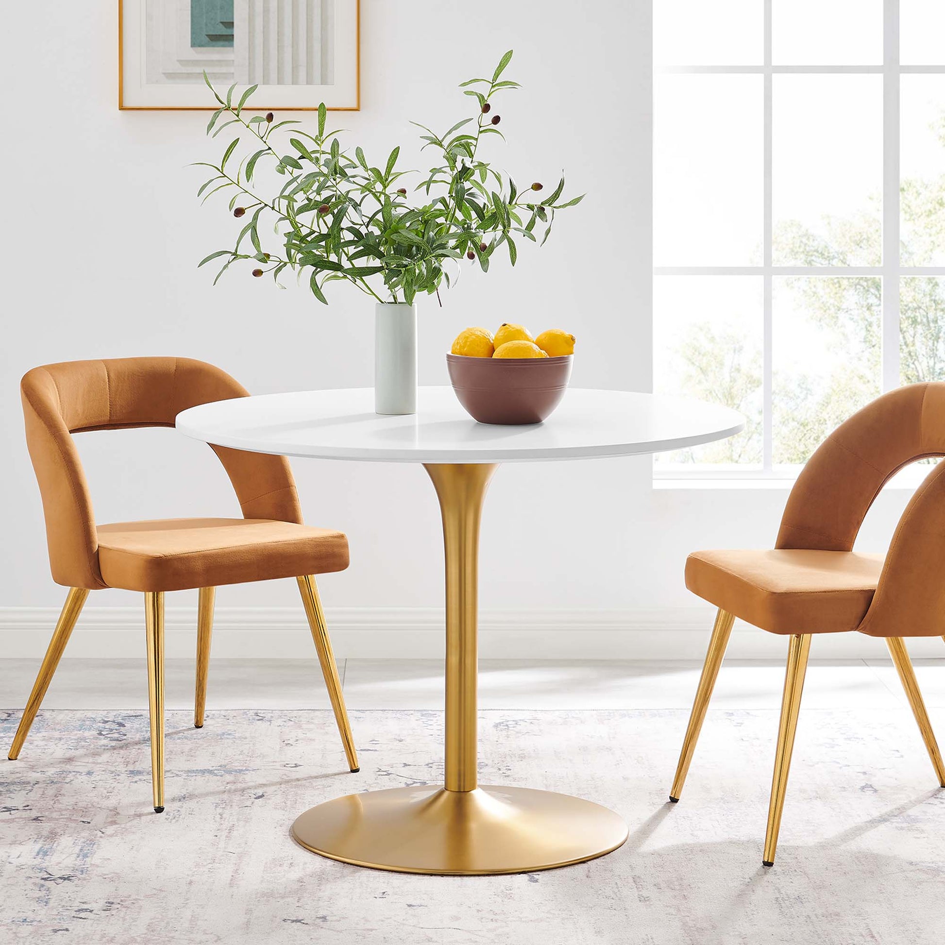 White round dining table with gold pedestal base, orange upholstered chairs, vase of greenery, lemons, sunlit dining nook.