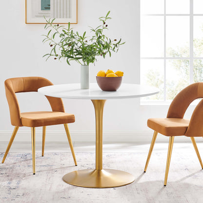 White round dining table with gold pedestal base, orange upholstered chairs, vase of greenery, lemons, sunlit dining nook.