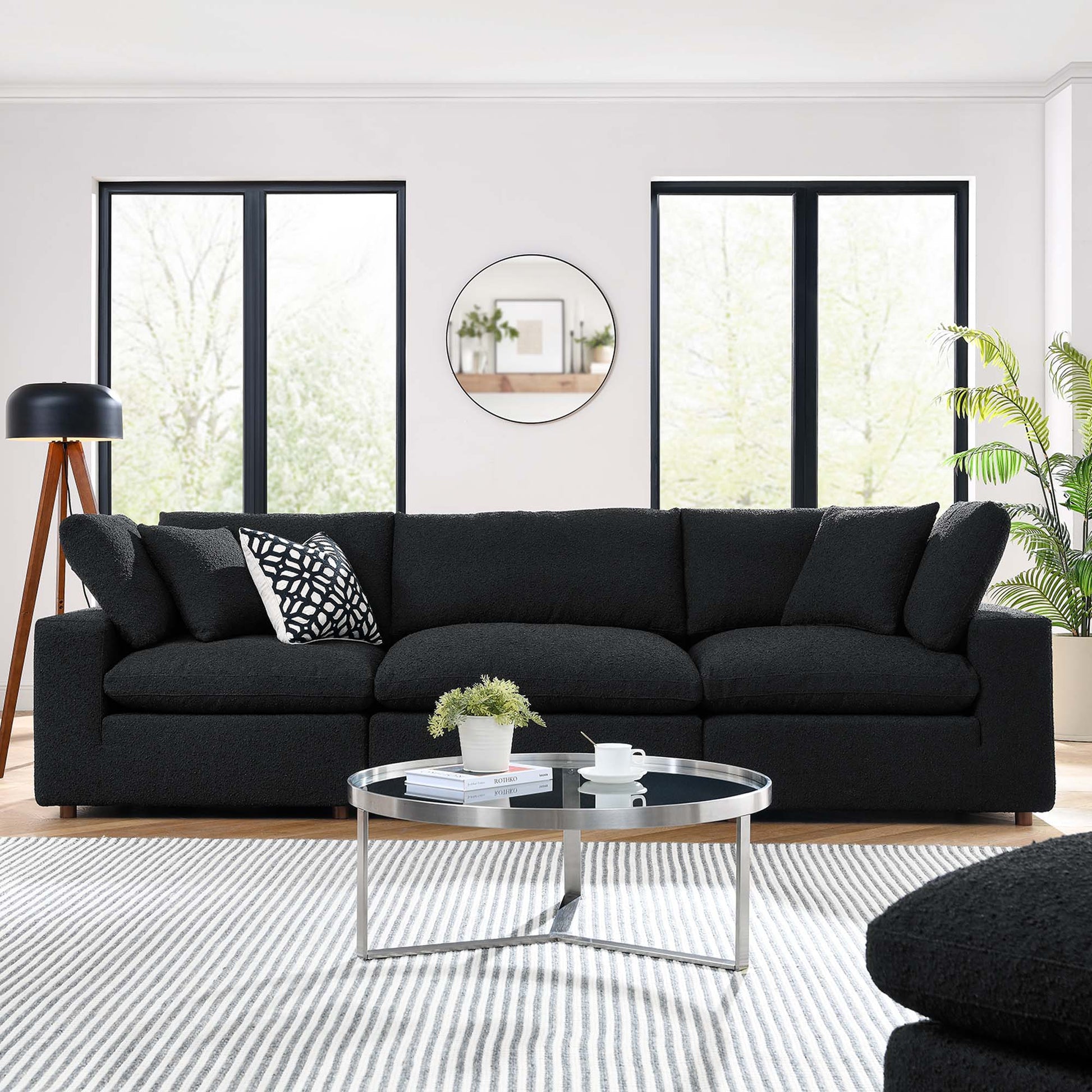 Black boucle fabric 3-seater sofa with cushions, round glass coffee table on a striped rug in a bright modern living room.