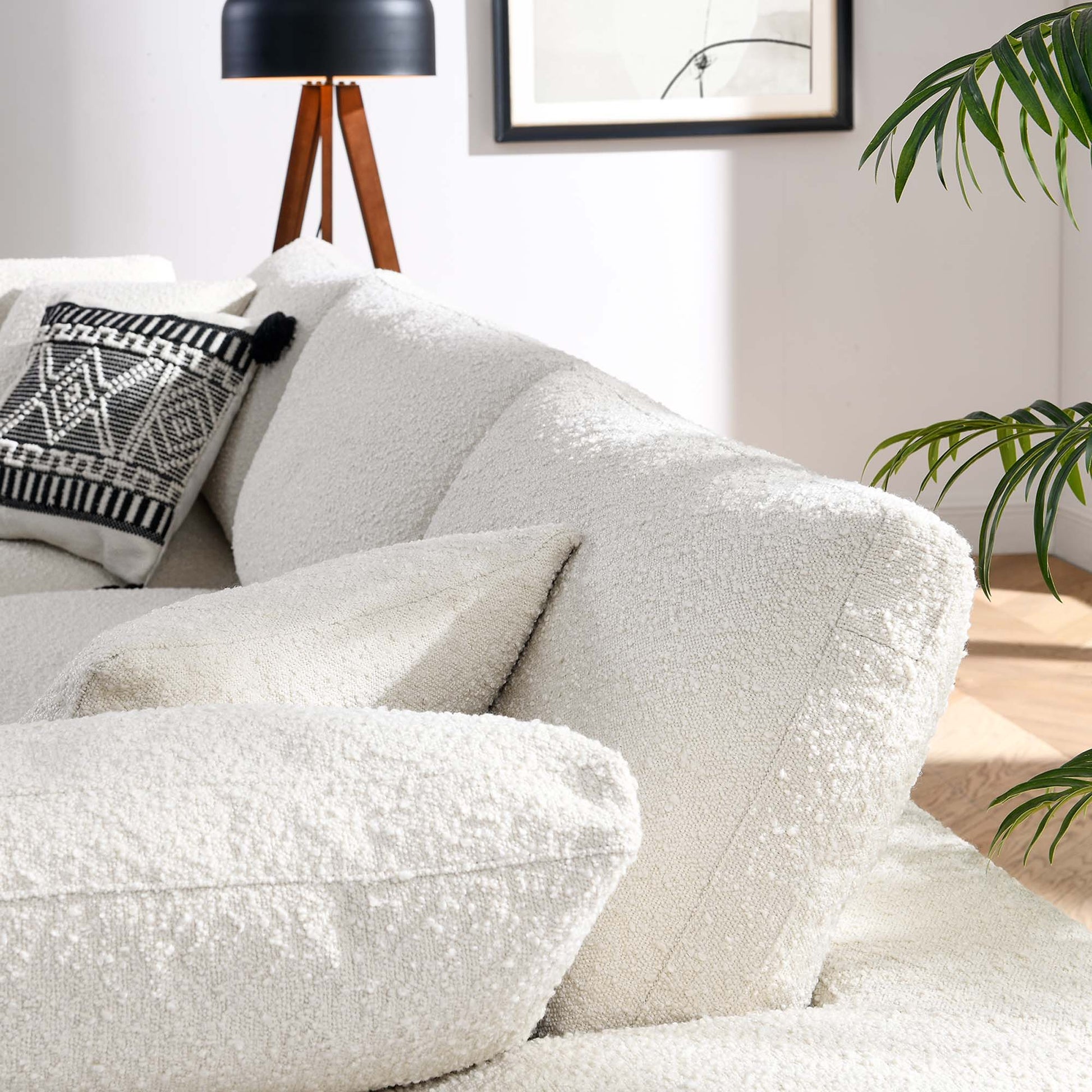 Ivory boucle sofa with plush cushions, black-and-white geometric pillow; tripod wooden lamp, framed art, and a green plant in a modern living room.