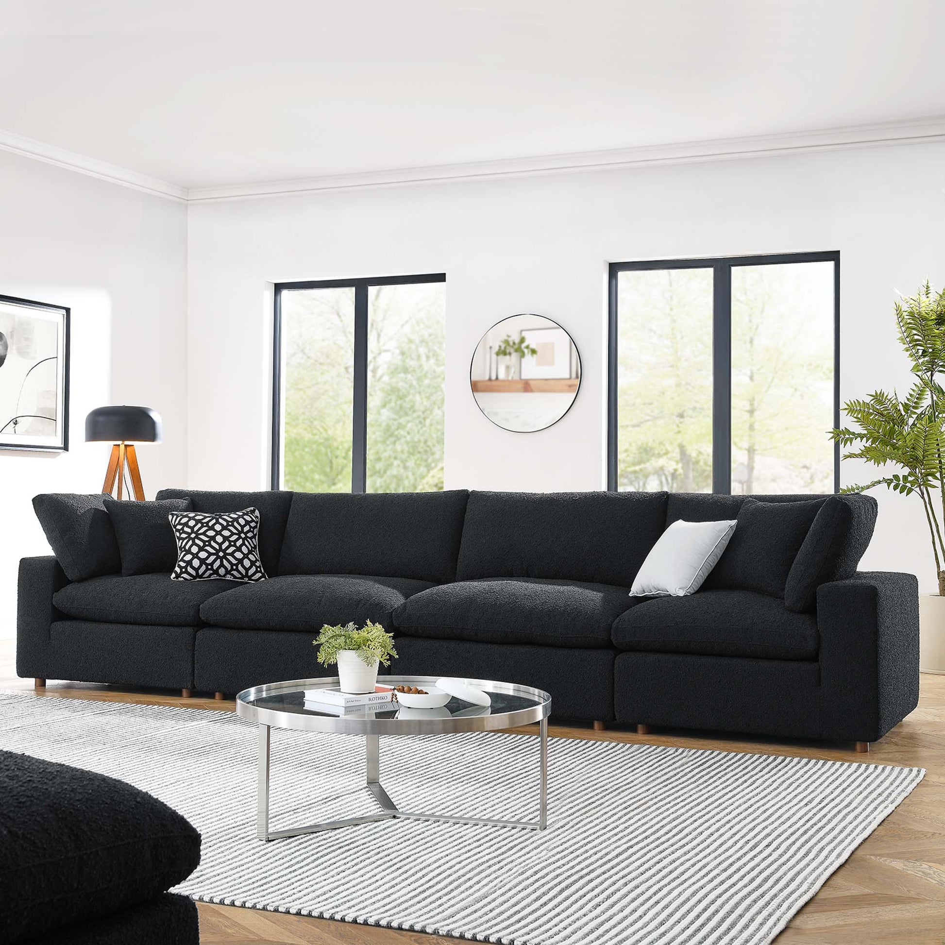 Modern black bouclé fabric 4-seater sofa with light wood legs, round glass coffee table, striped rug, tripod lamp in a bright living room.