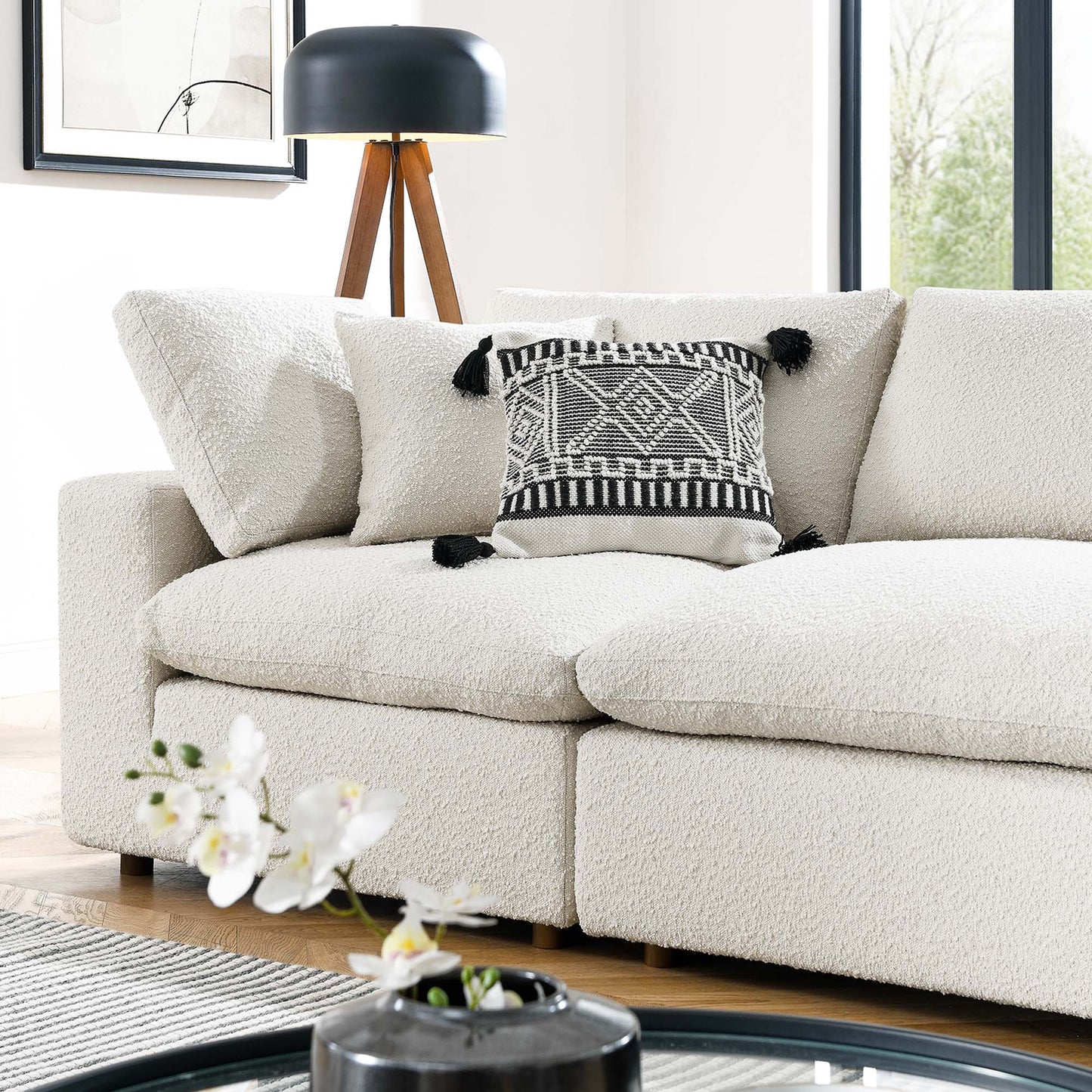 Ivory boucle sofa with a geometric black & white pillow with tassels and a black tripod floor lamp in a bright living room.